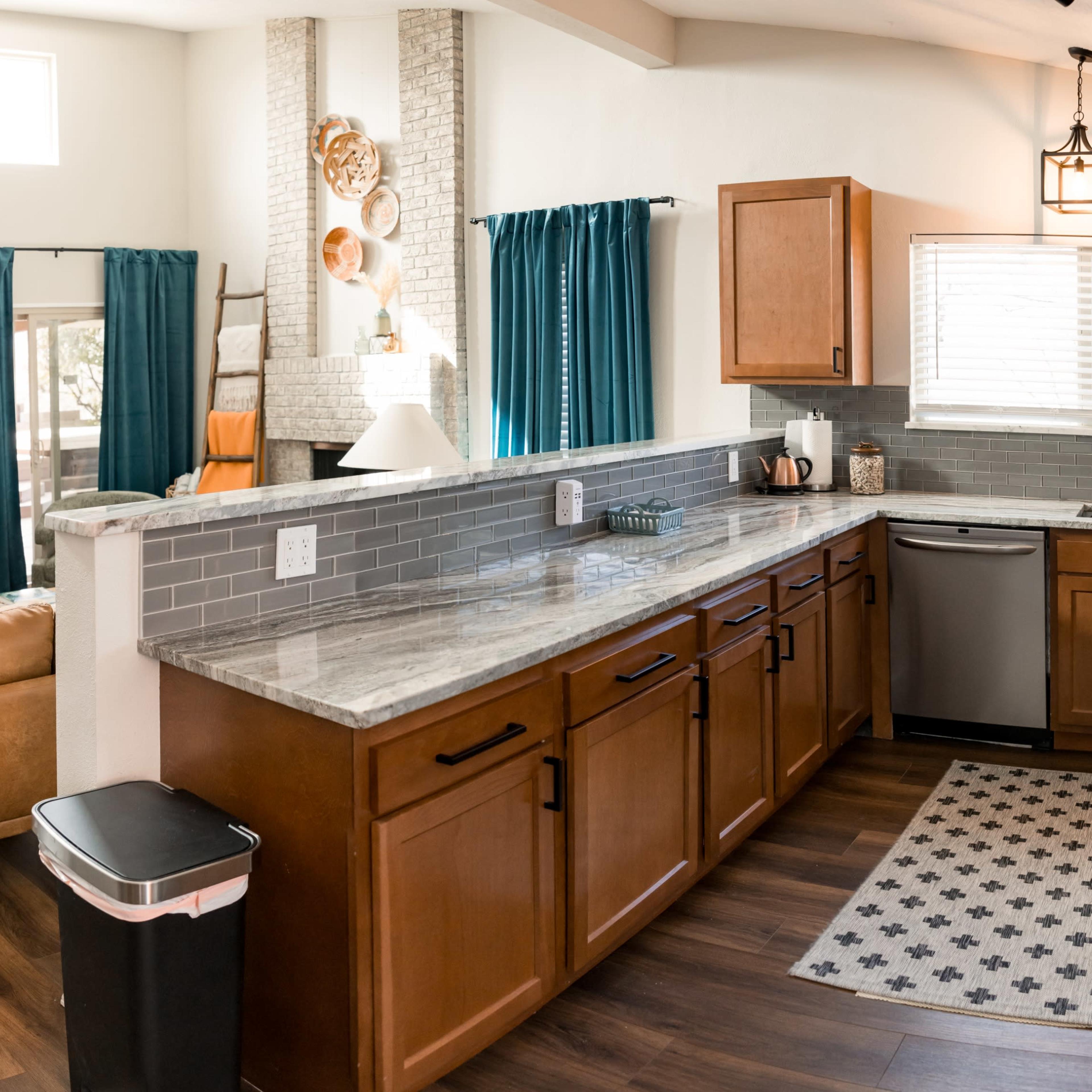 A modern kitchen with wooden cabinets, a stainless steel dishwasher, and a view of an open living area featuring a couch and a decorative fireplace.