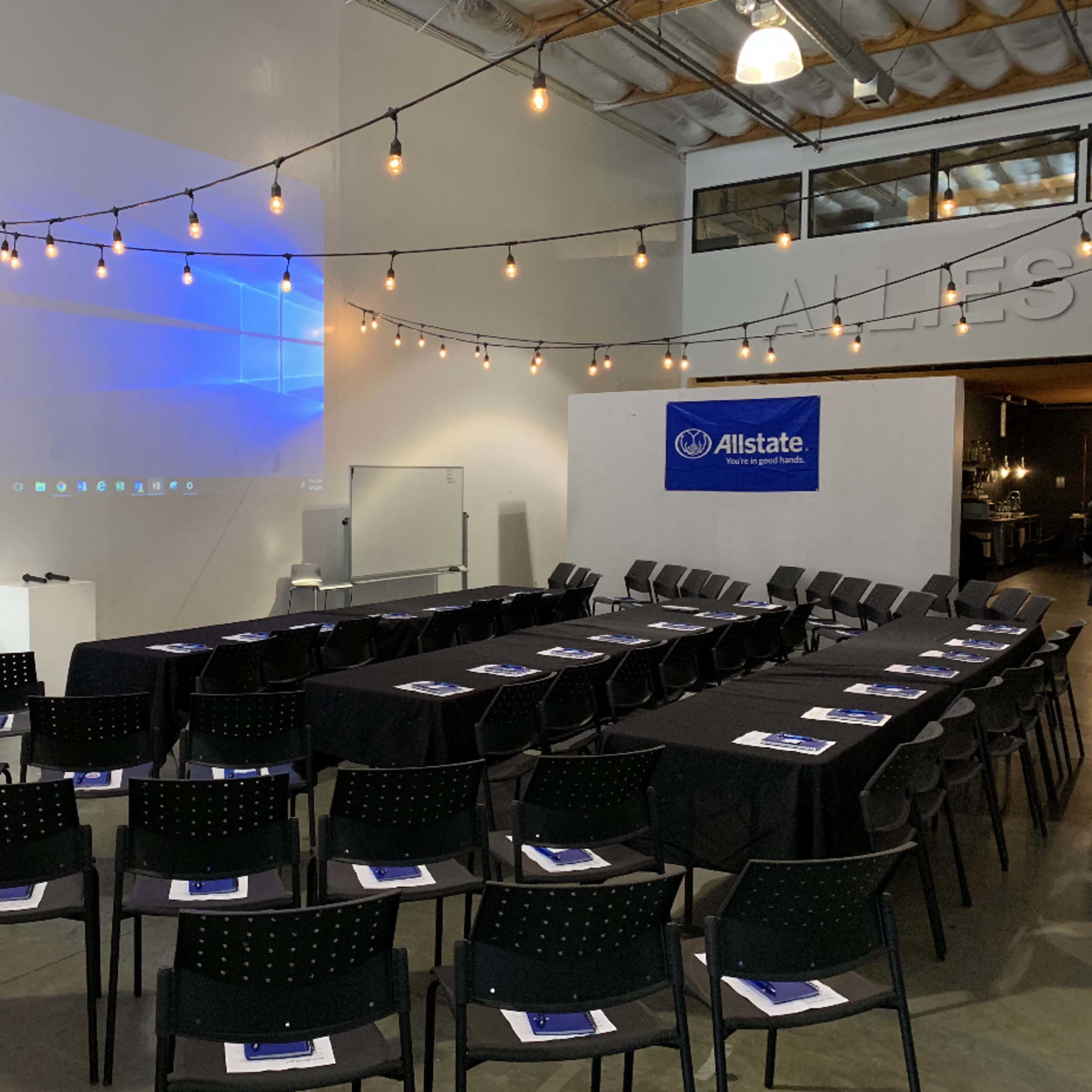 A spacious meeting room is set up with rows of chairs facing a projector screen, decorated with string lights above.