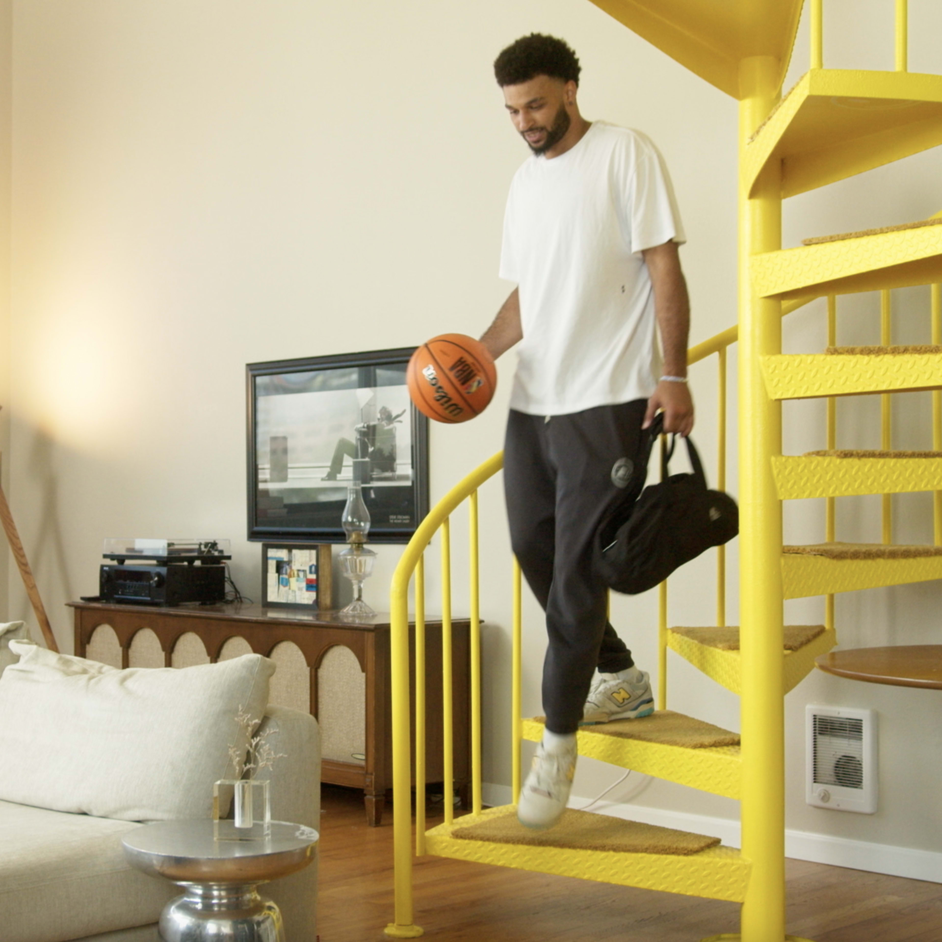 A man descends a yellow spiral staircase while holding a basketball and a black gym bag in a living room with a modern aesthetic.