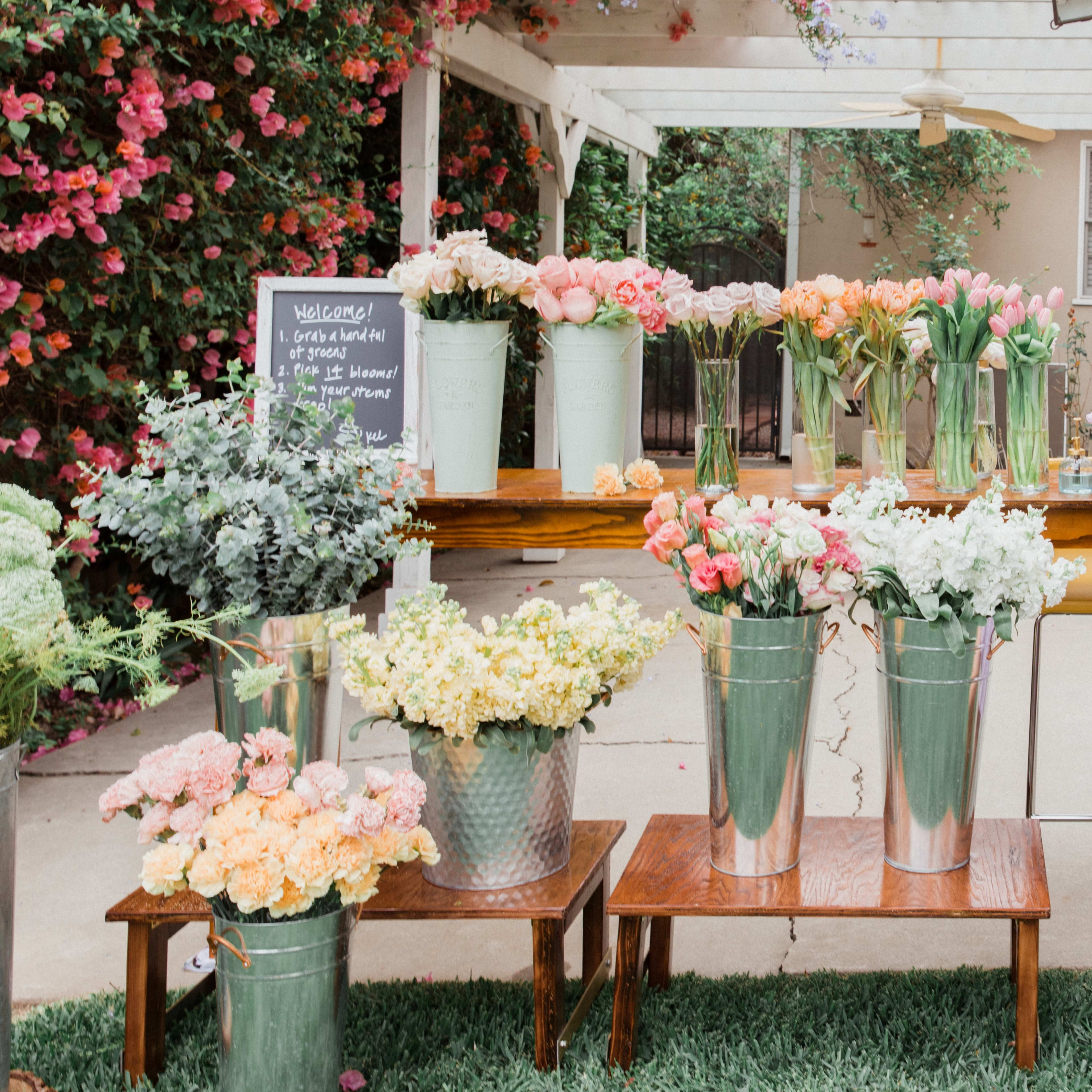 A display of various flowers in metal buckets is arranged on wooden tables under a white gazebo adorned with colorful blooms.