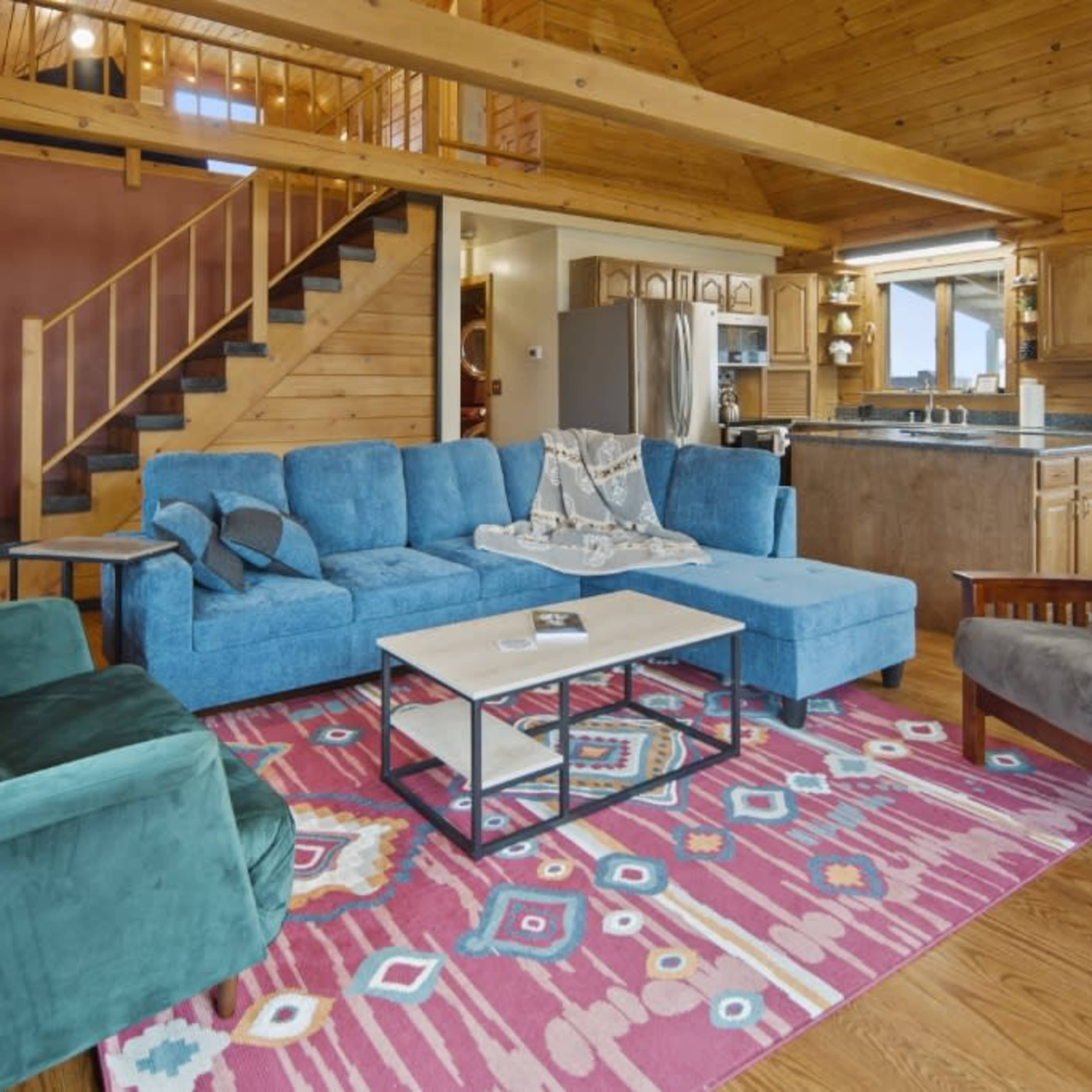 The room features a spacious living area with a sectional sofa, a coffee table, and a colorful area rug, all set against a backdrop of wooden walls and a staircase leading to an upper level.