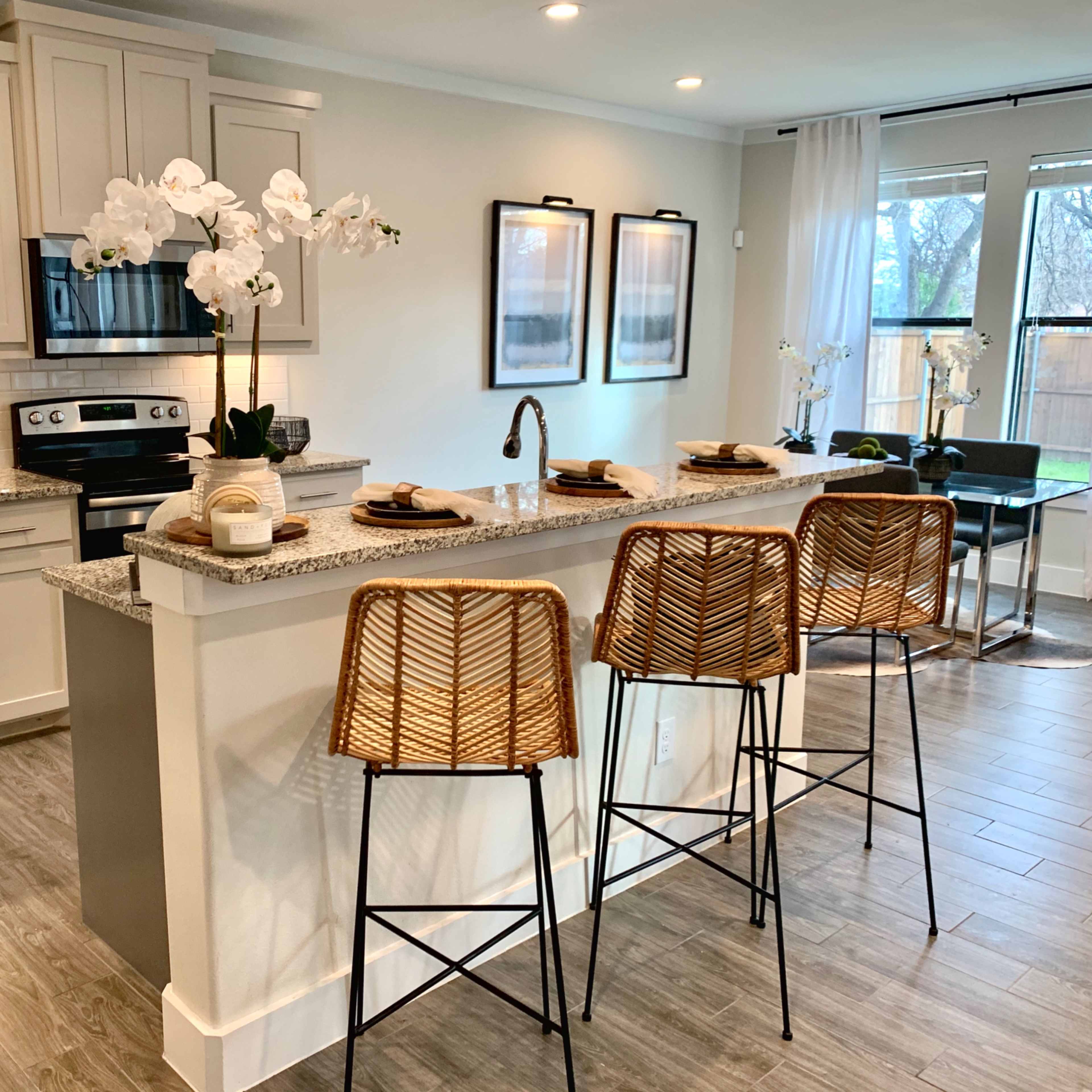 A modern kitchen features a central island with three rattan bar stools, accompanied by stainless steel appliances and decorative plants.