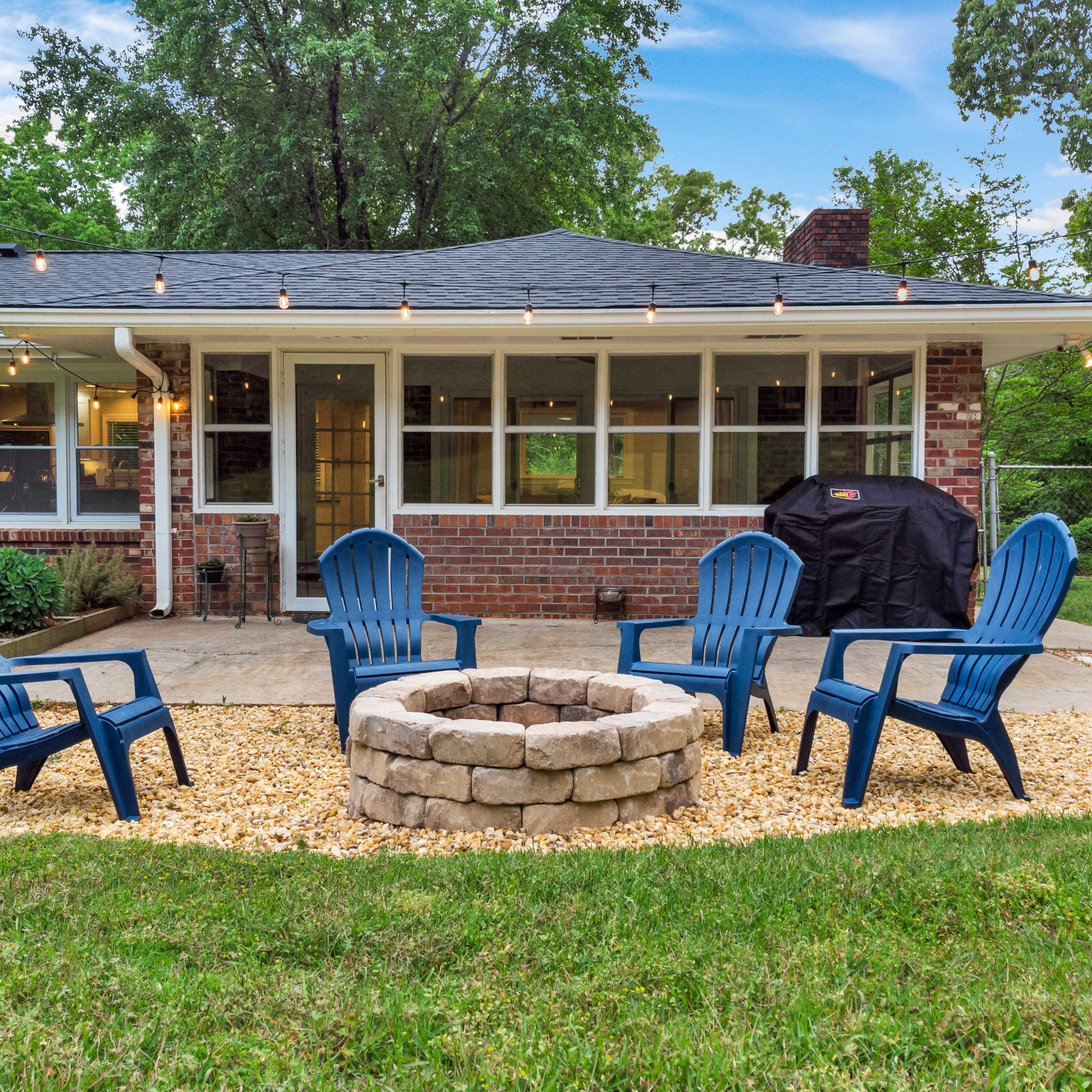 A backyard patio setup with four blue Adirondack chairs arranged around a stone fire pit, surrounded by a gravel area and illuminated by string lights.