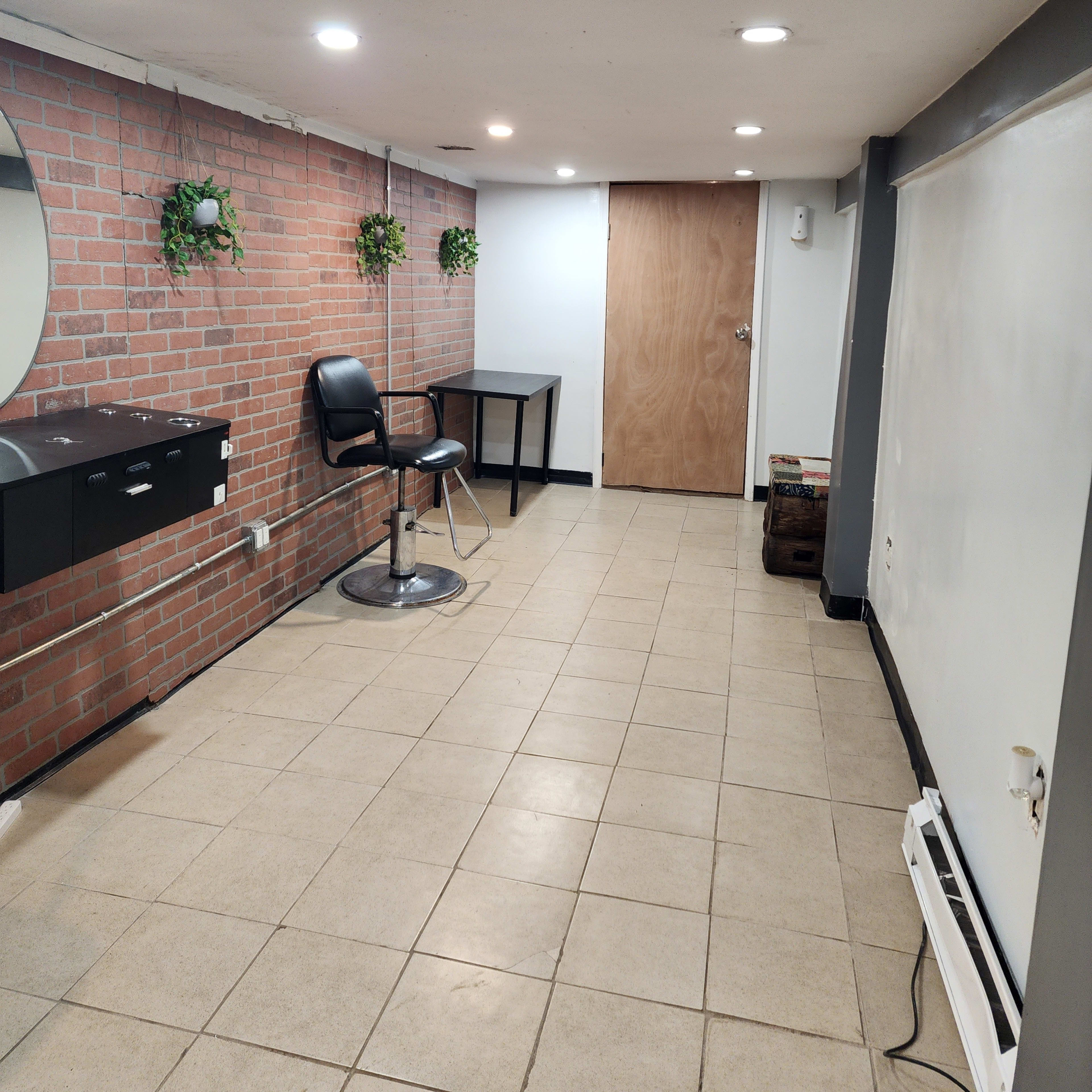 A clean, empty room with a brick wall, a large mirror, a black chair, a small table, and a door at one end.