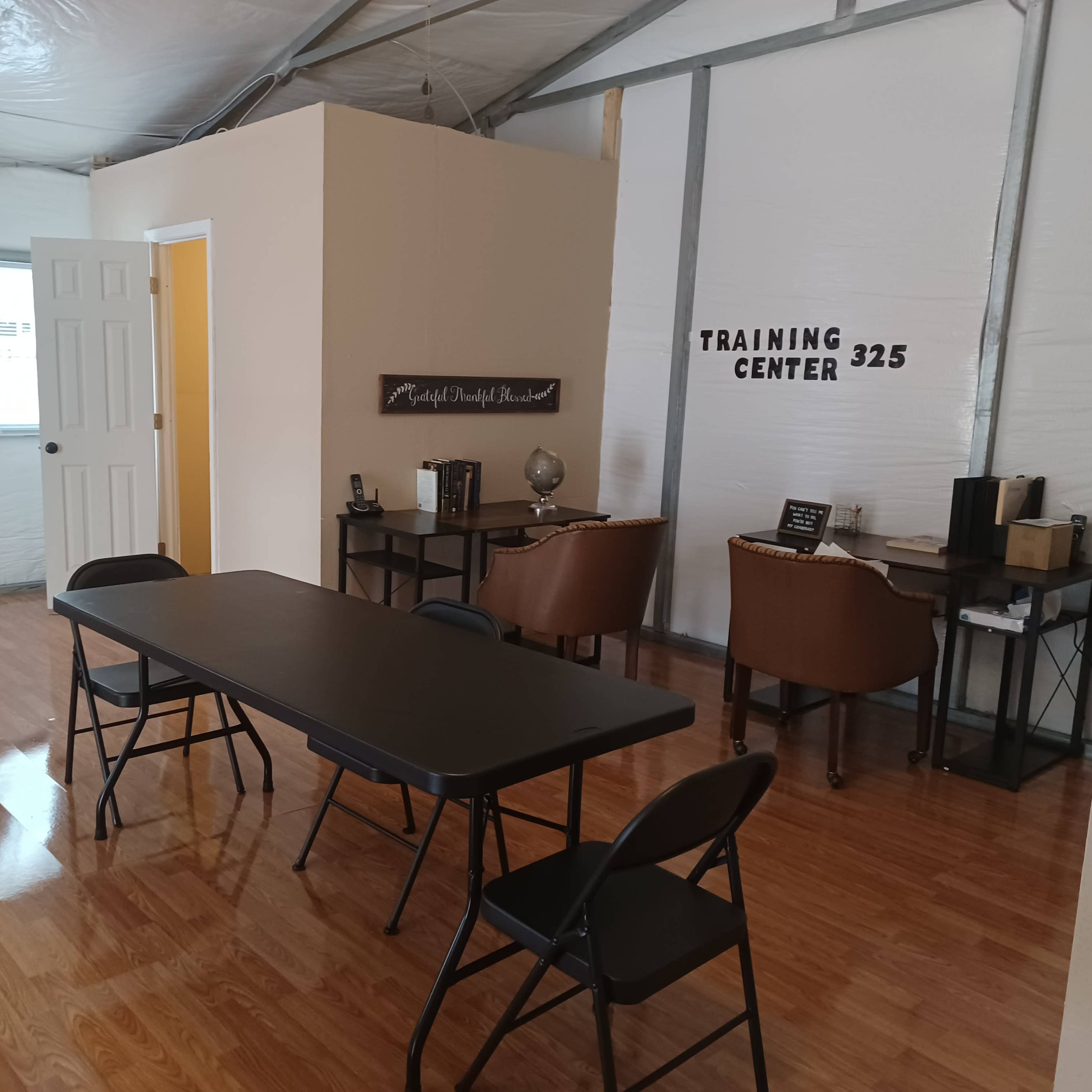An interior of a training center featuring a table with chairs, a door leading to another room, and two chairs beside a desk.