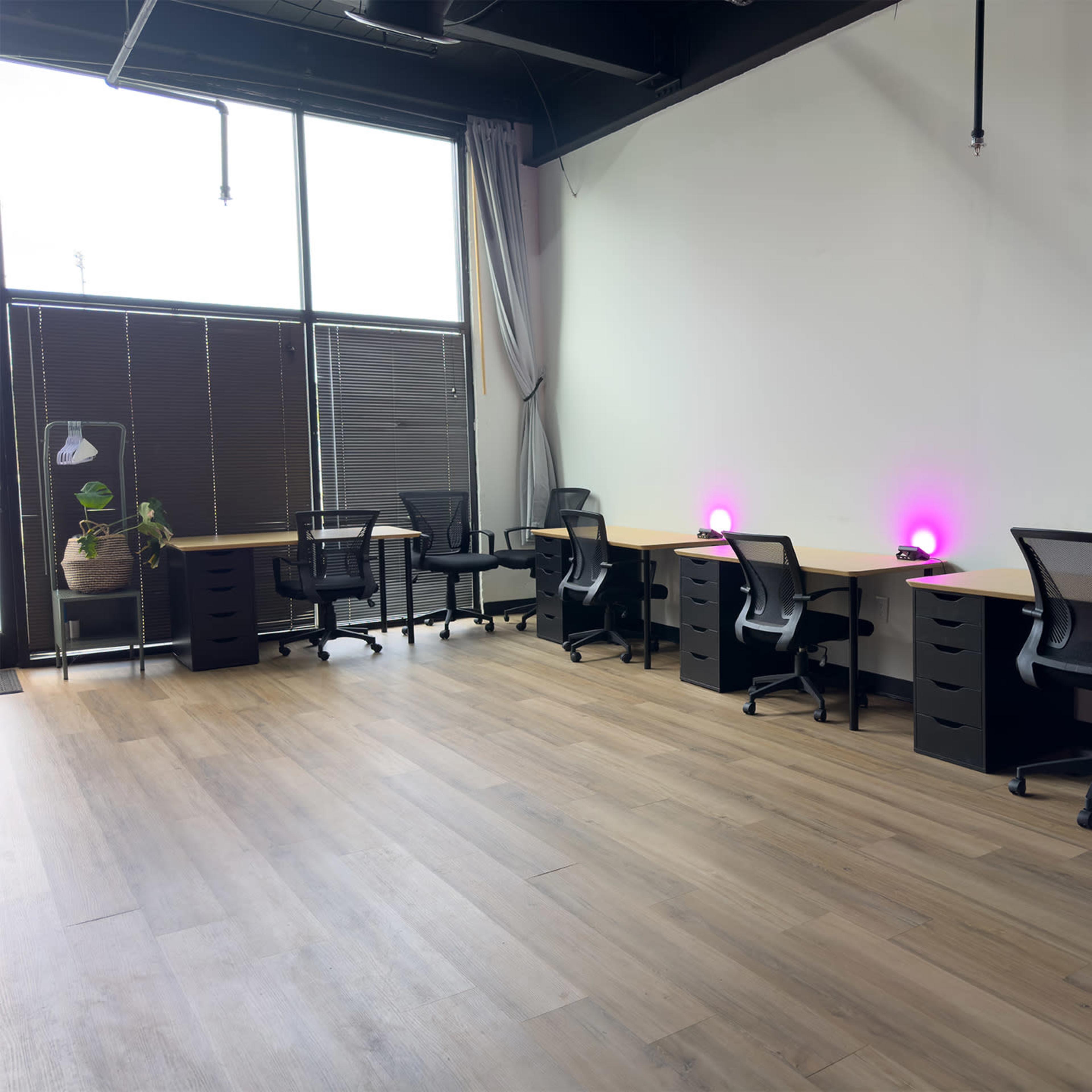 A coworking space with several desks and ergonomic chairs, illuminated by purple lights, along with large windows letting in natural light.