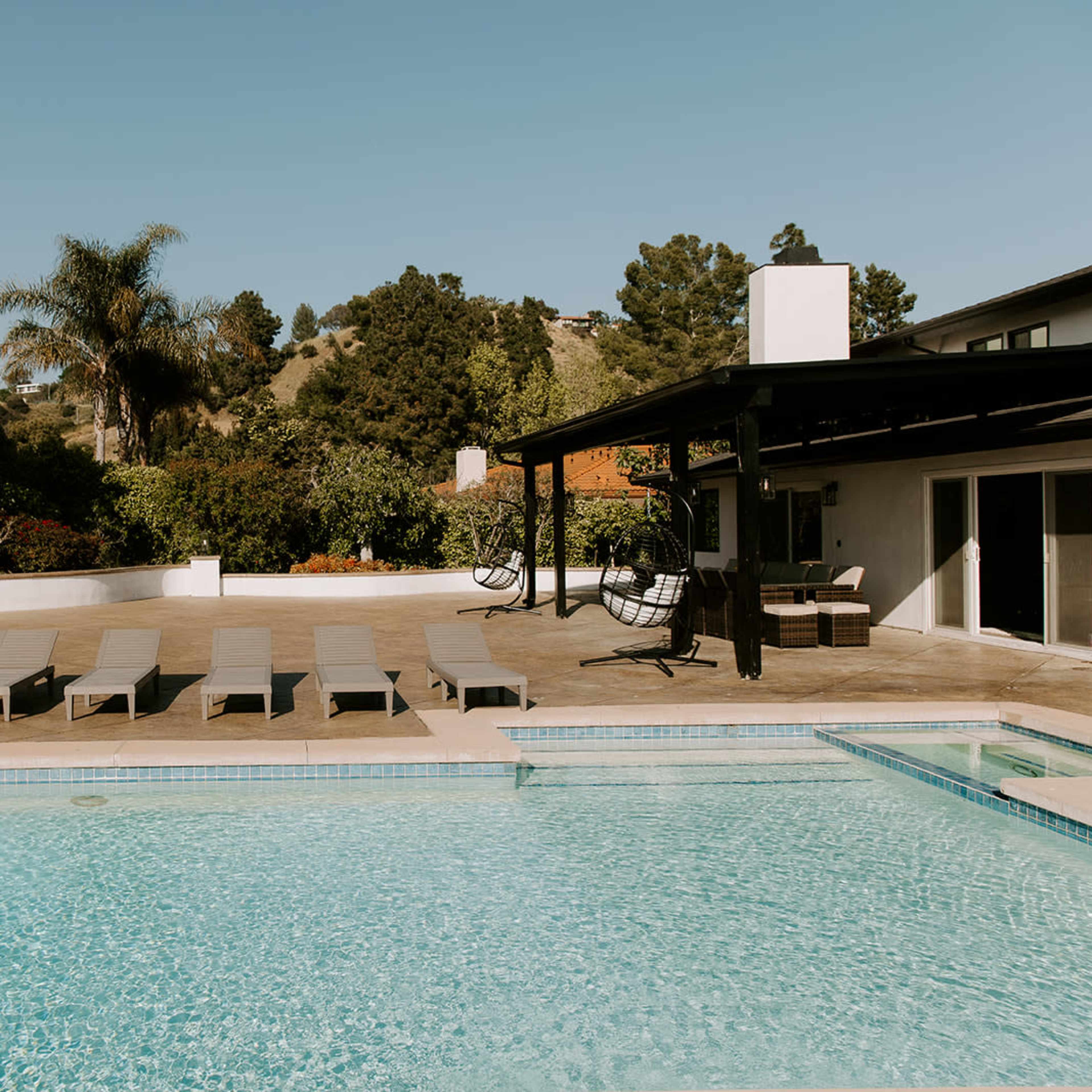 A clear swimming pool is surrounded by lounge chairs and a shaded seating area, with trees and hills in the background.