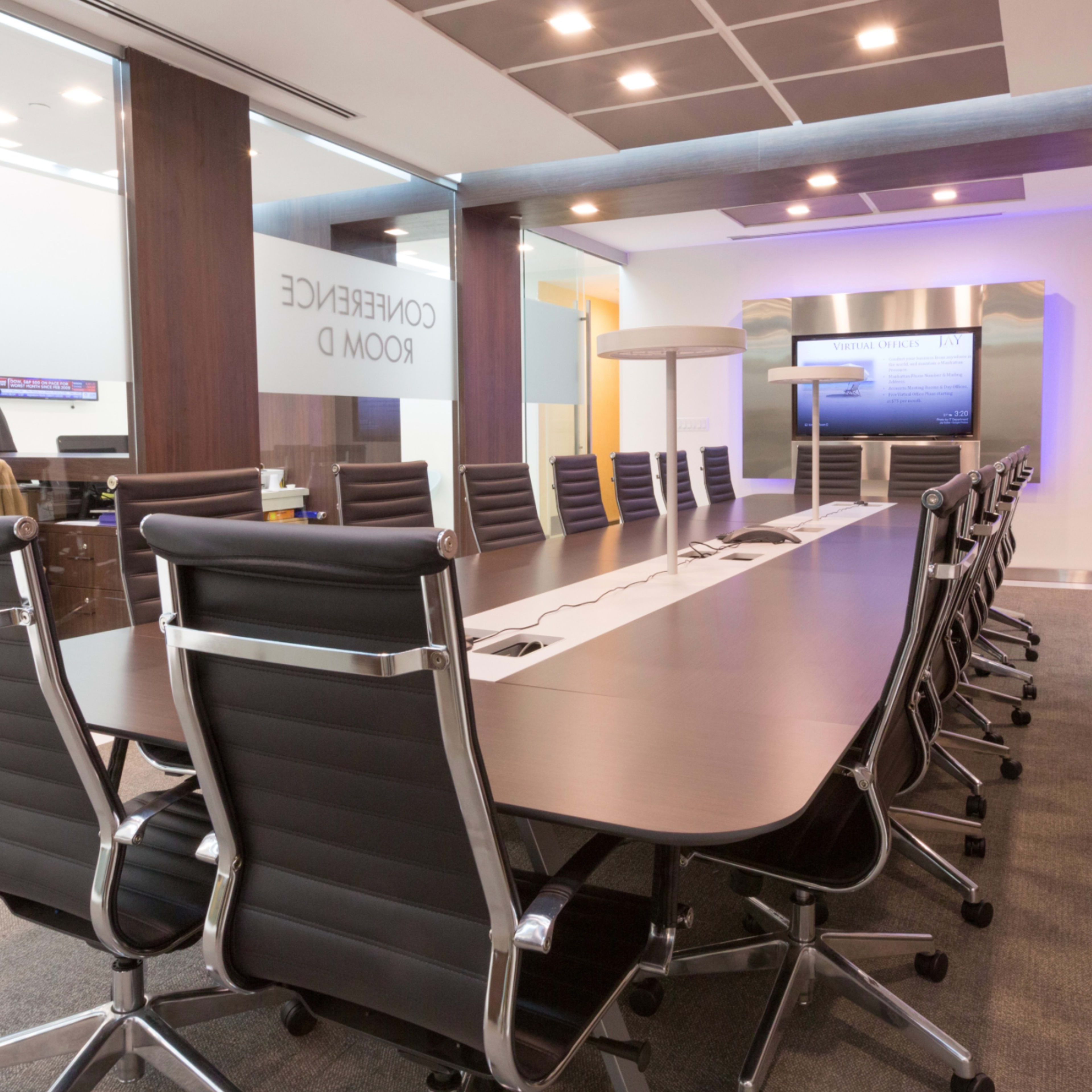 A large conference room features a long table surrounded by black leather chairs, with modern decor and a presentation screen at one end.