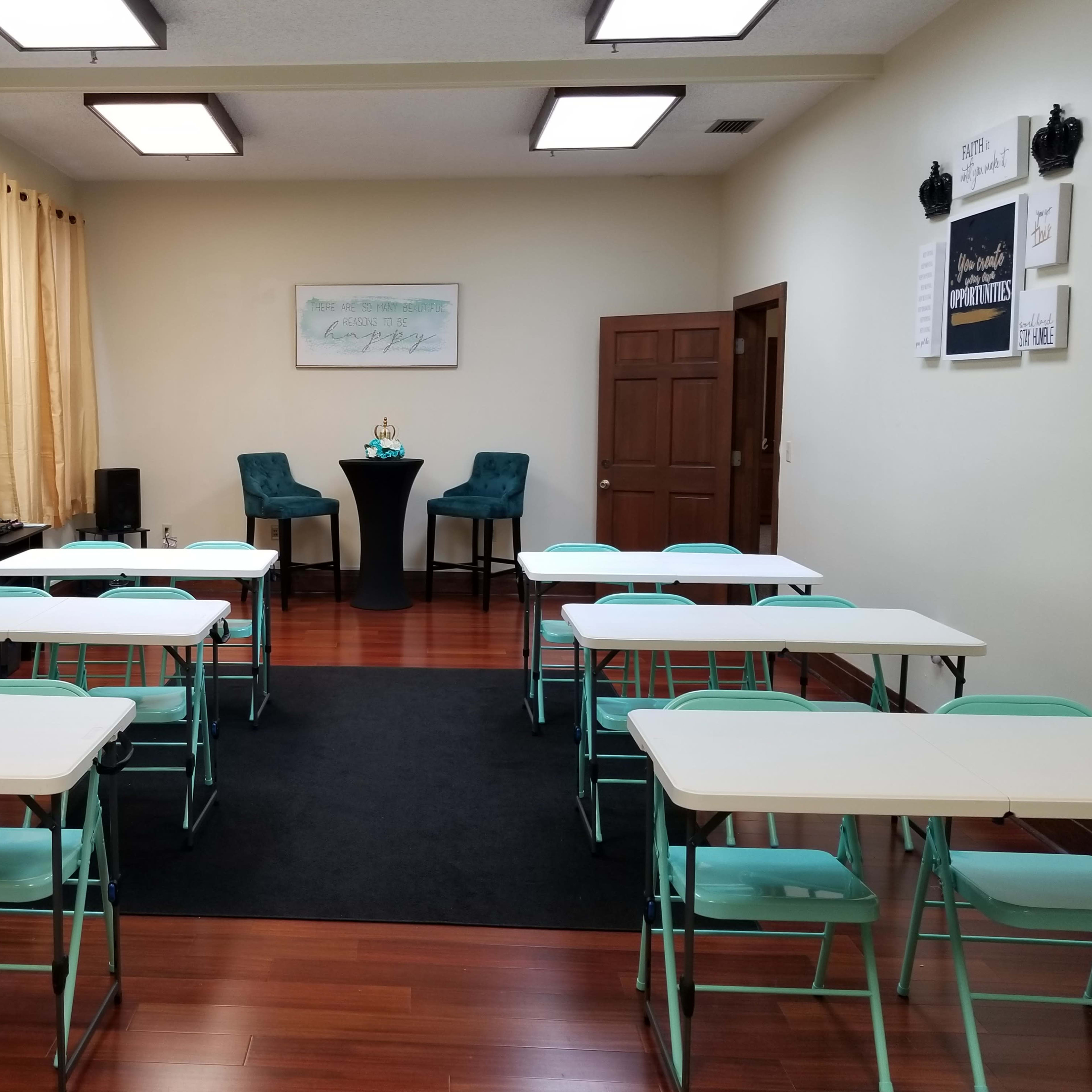 The room features several rows of white tables and teal chairs, with a small table set up at the front and large windows adorned with yellow curtains.