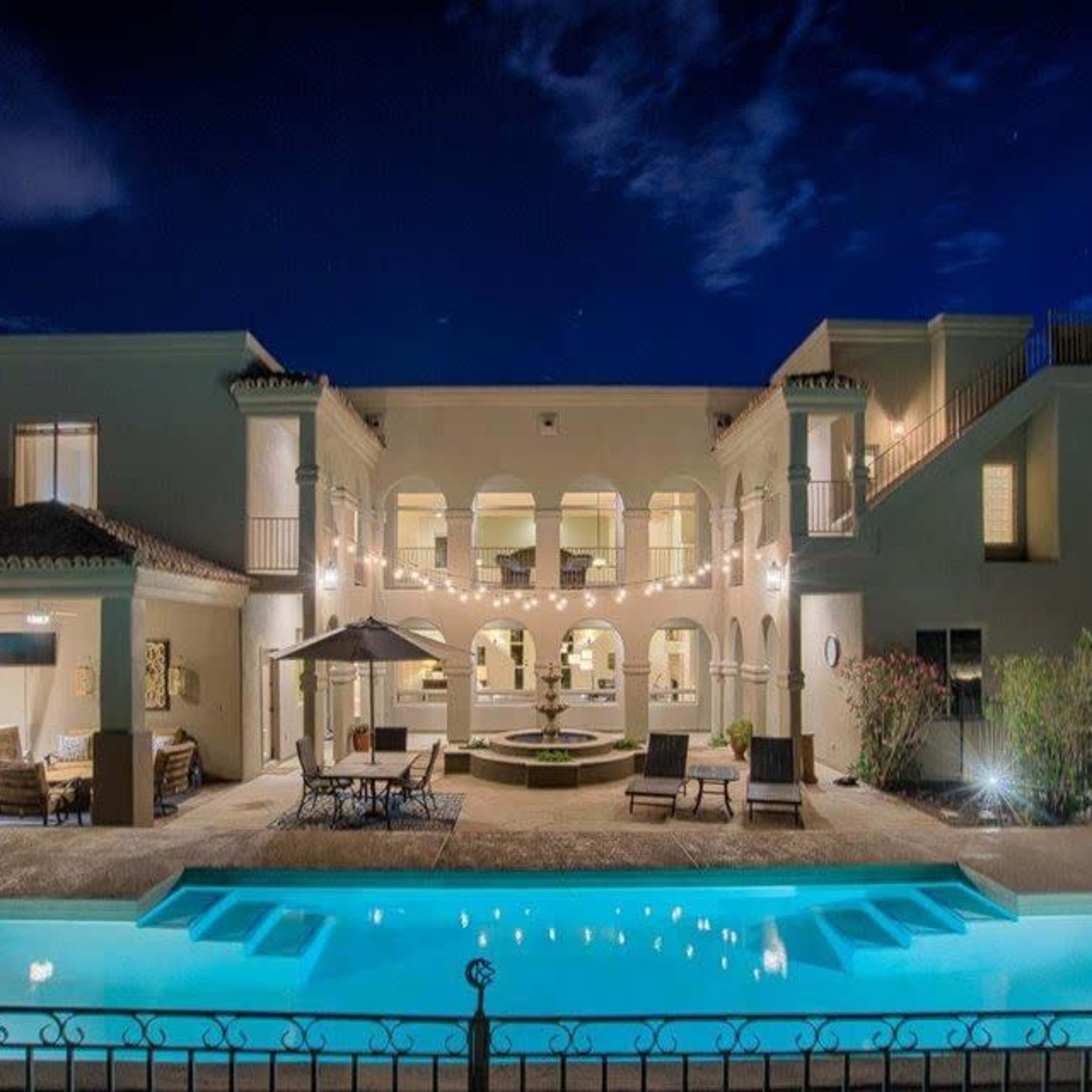 A large, illuminated house with multiple arches, a swimming pool in the foreground, and outdoor seating areas under a clear night sky.