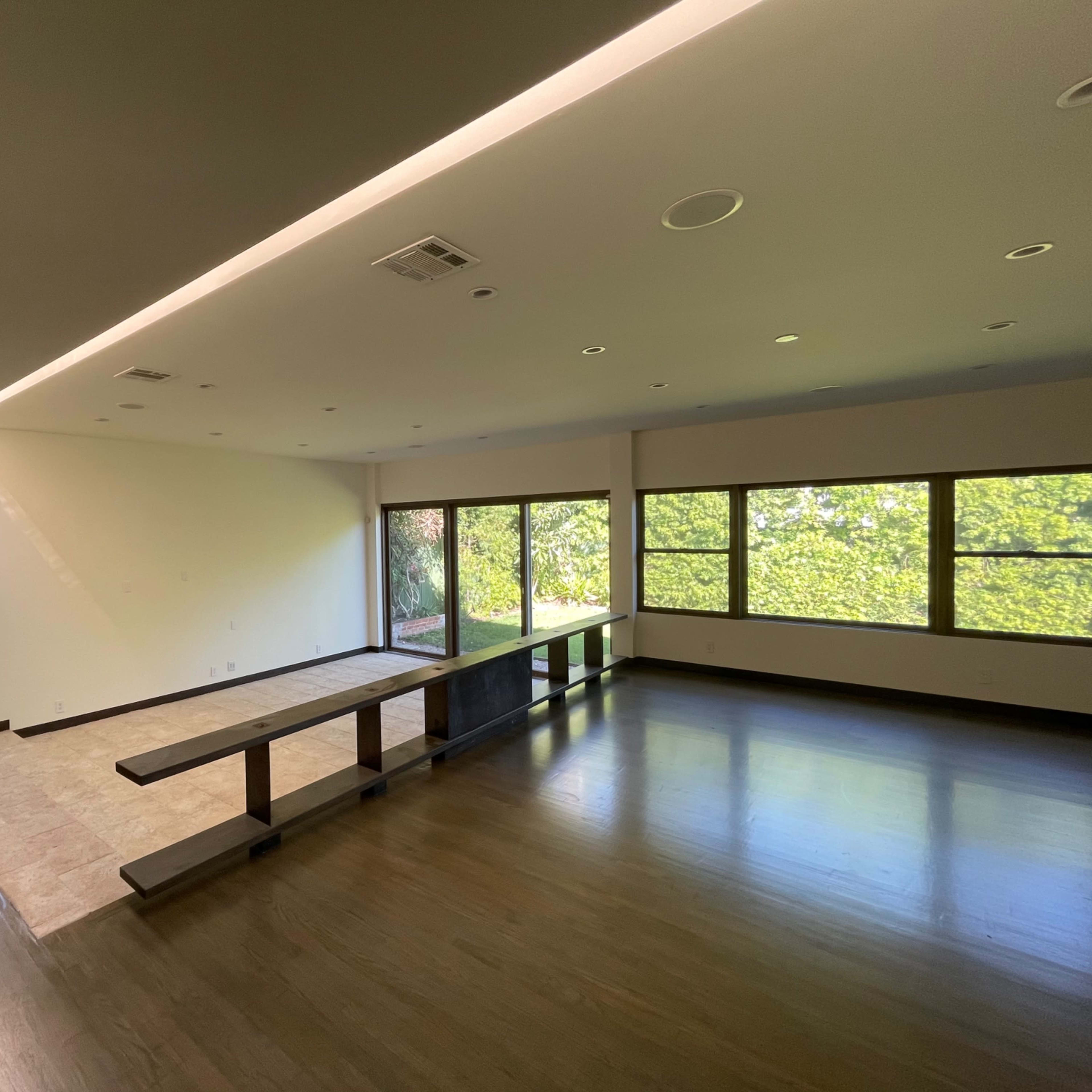 A spacious room with large windows overlooking greenery, featuring a long table in the center and a mix of wood and tile flooring.
