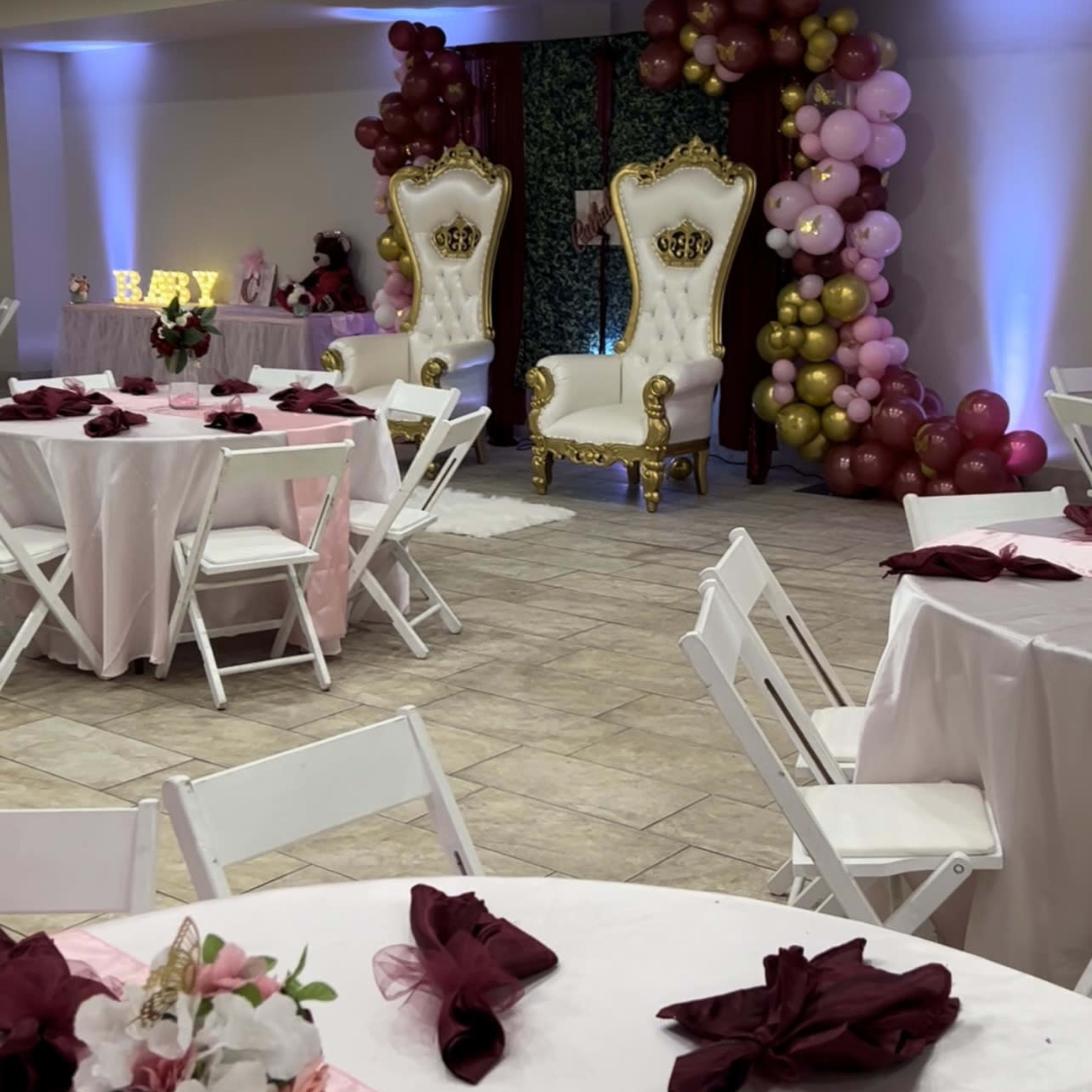 The scene features a decorated event space with white tables, maroon napkins, and two golden thrones surrounded by balloon arrangements in various colors.