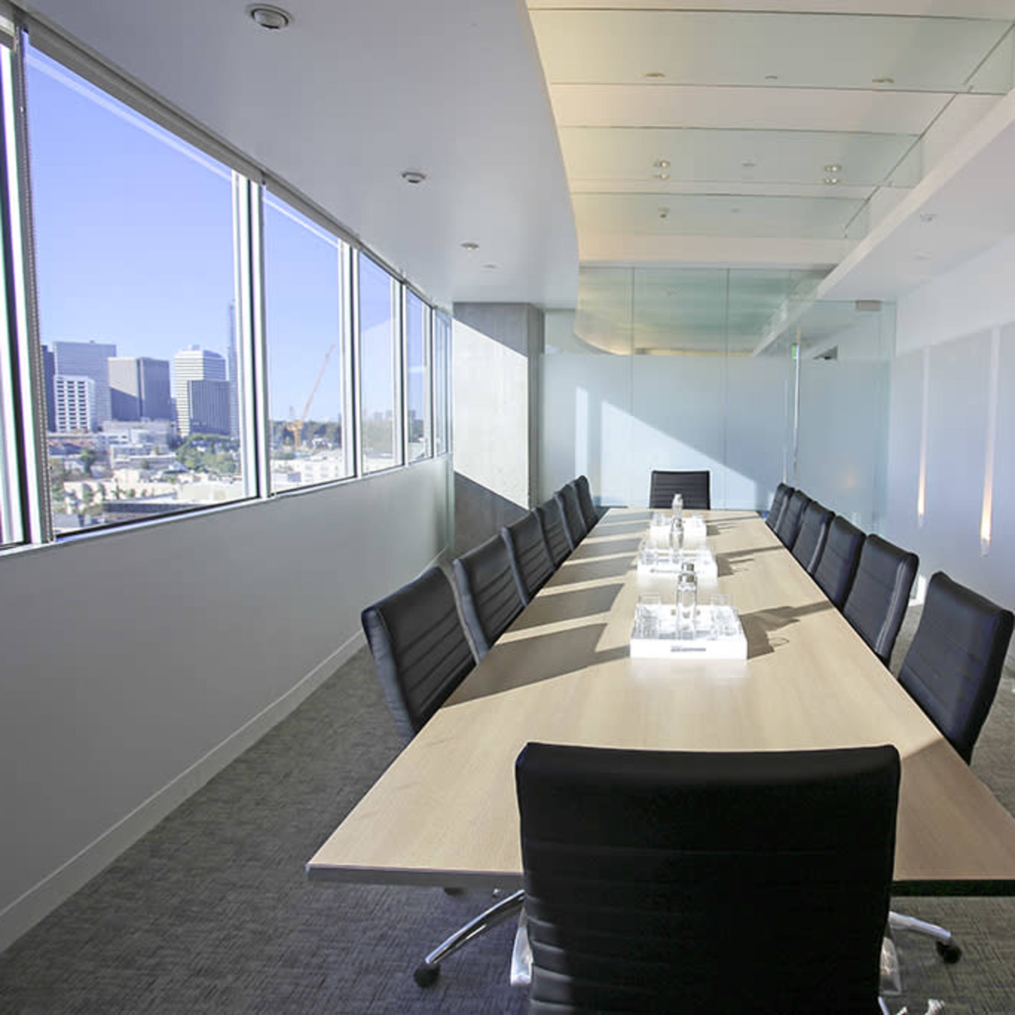 A modern conference room features a long wooden table surrounded by black chairs, with large windows offering a view of the city skyline.