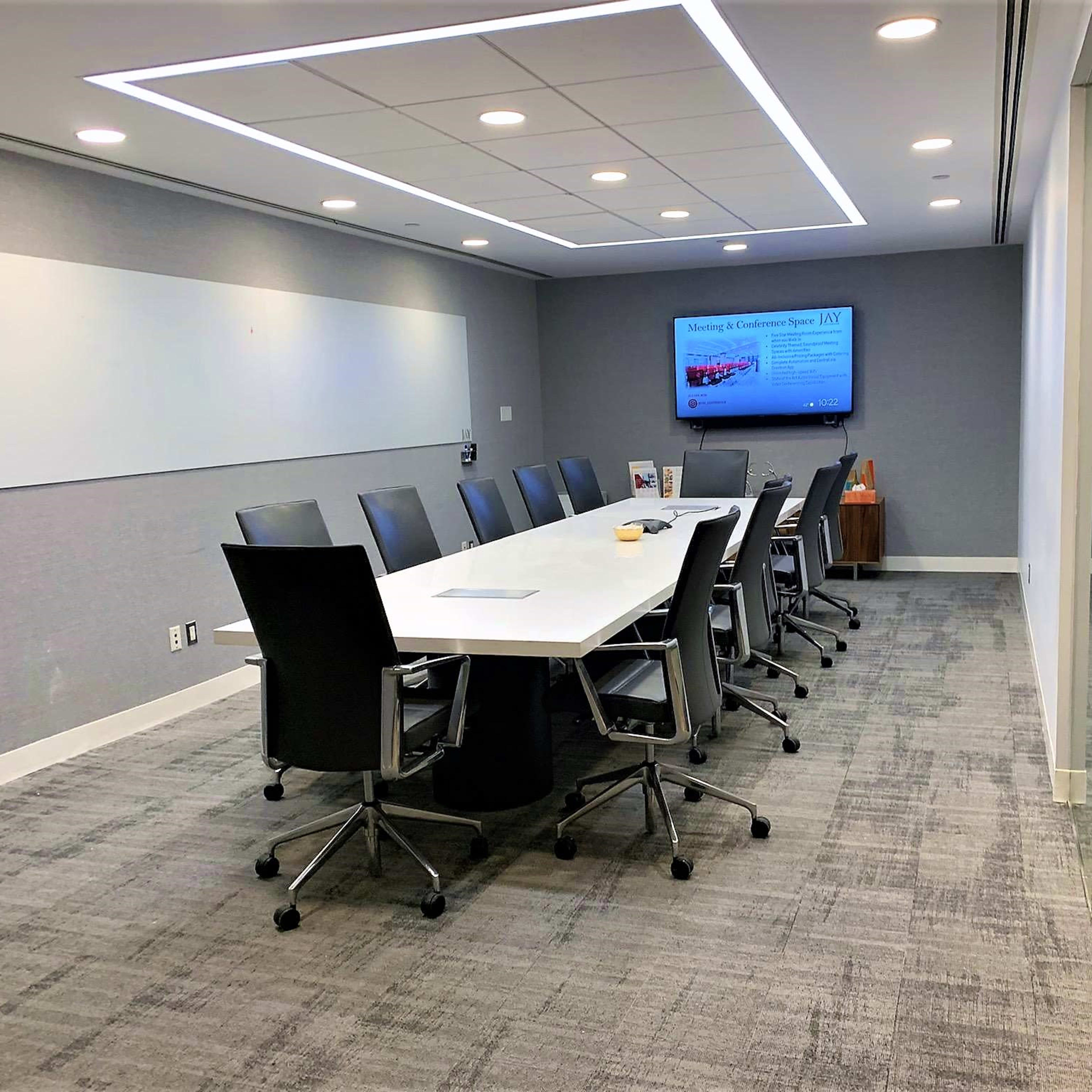 A modern conference room features a long white table surrounded by ergonomic black chairs, with a large screen displaying a presentation and a whiteboard on the wall.