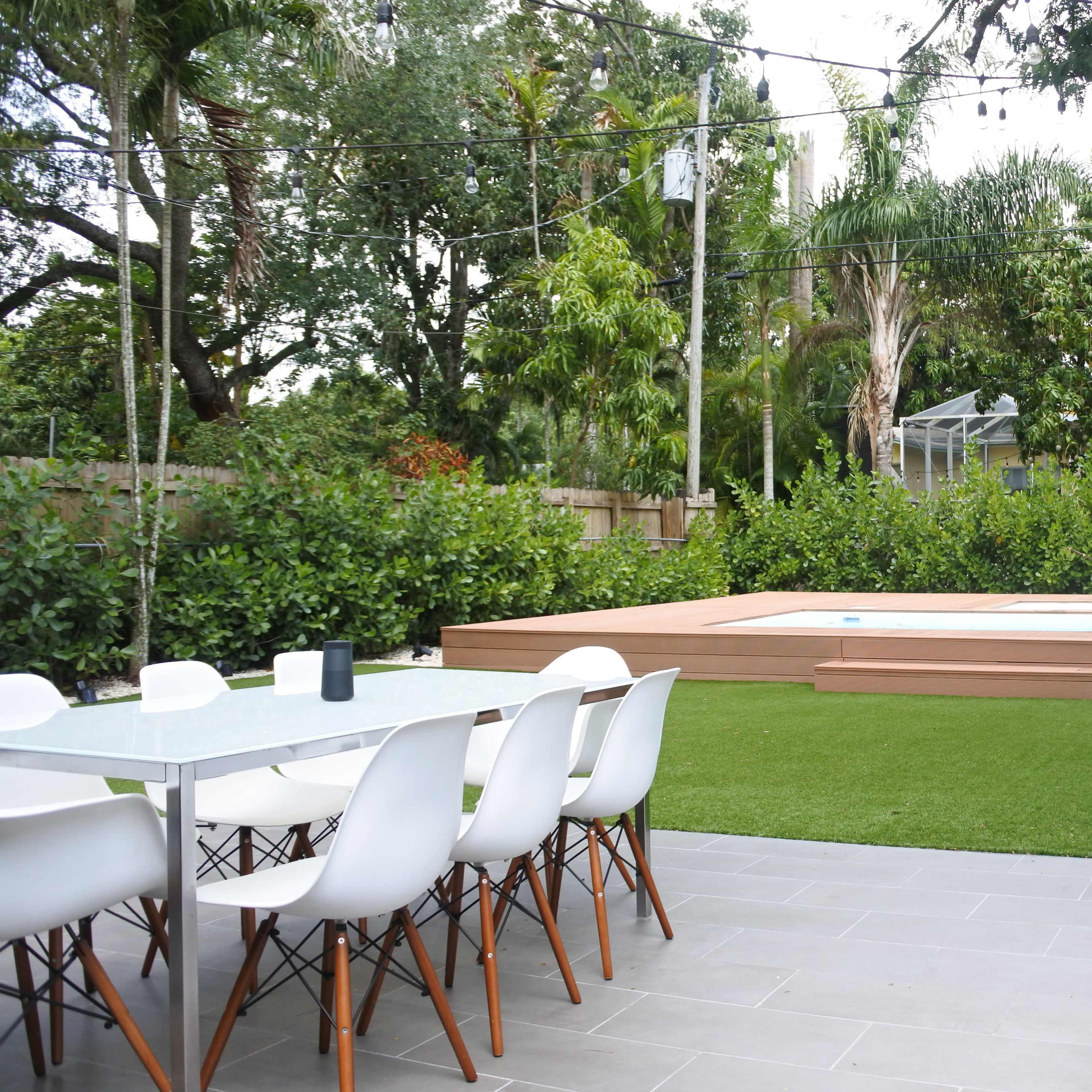 A modern outdoor patio features a dining table with white chairs, a small swimming pool, and lush greenery surrounding the area.