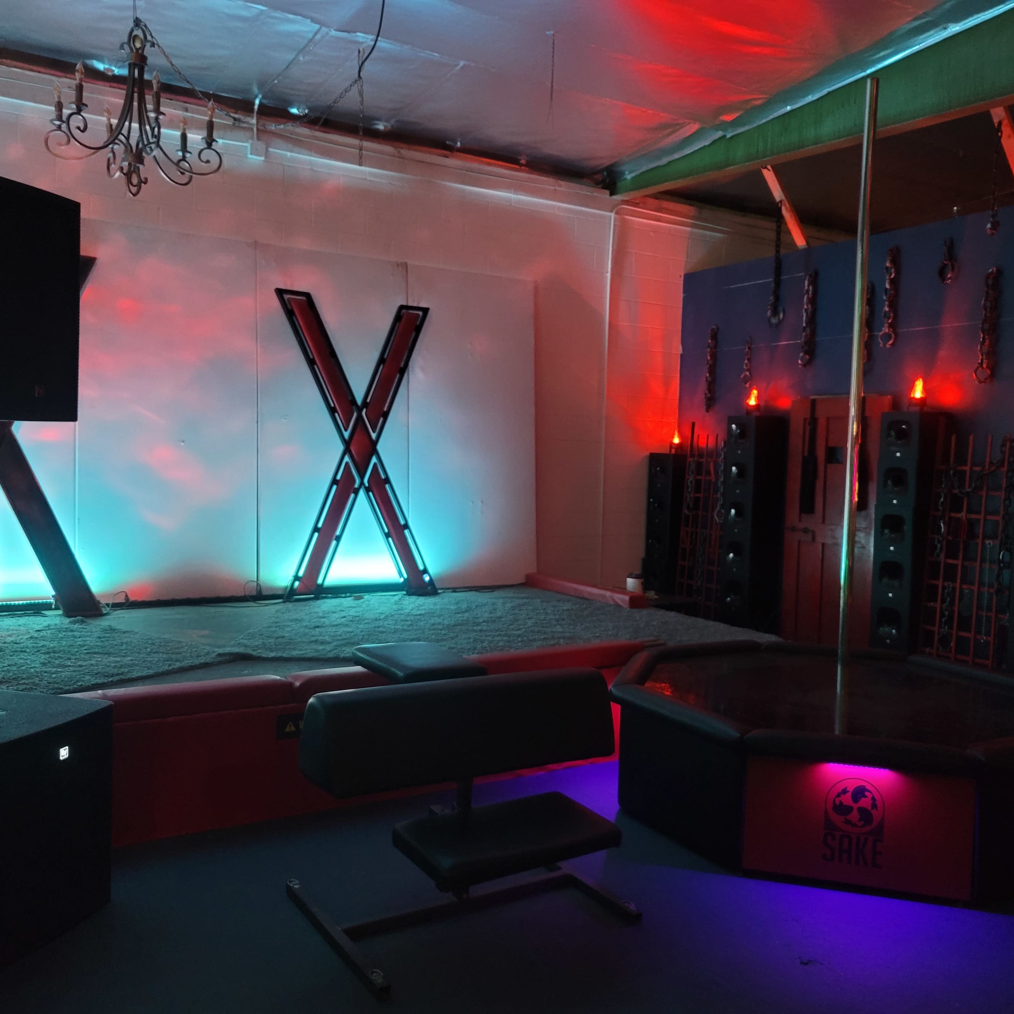 A dimly lit room designed for performances, featuring a pole in the center, large speakers along the walls, and decorative lighting.