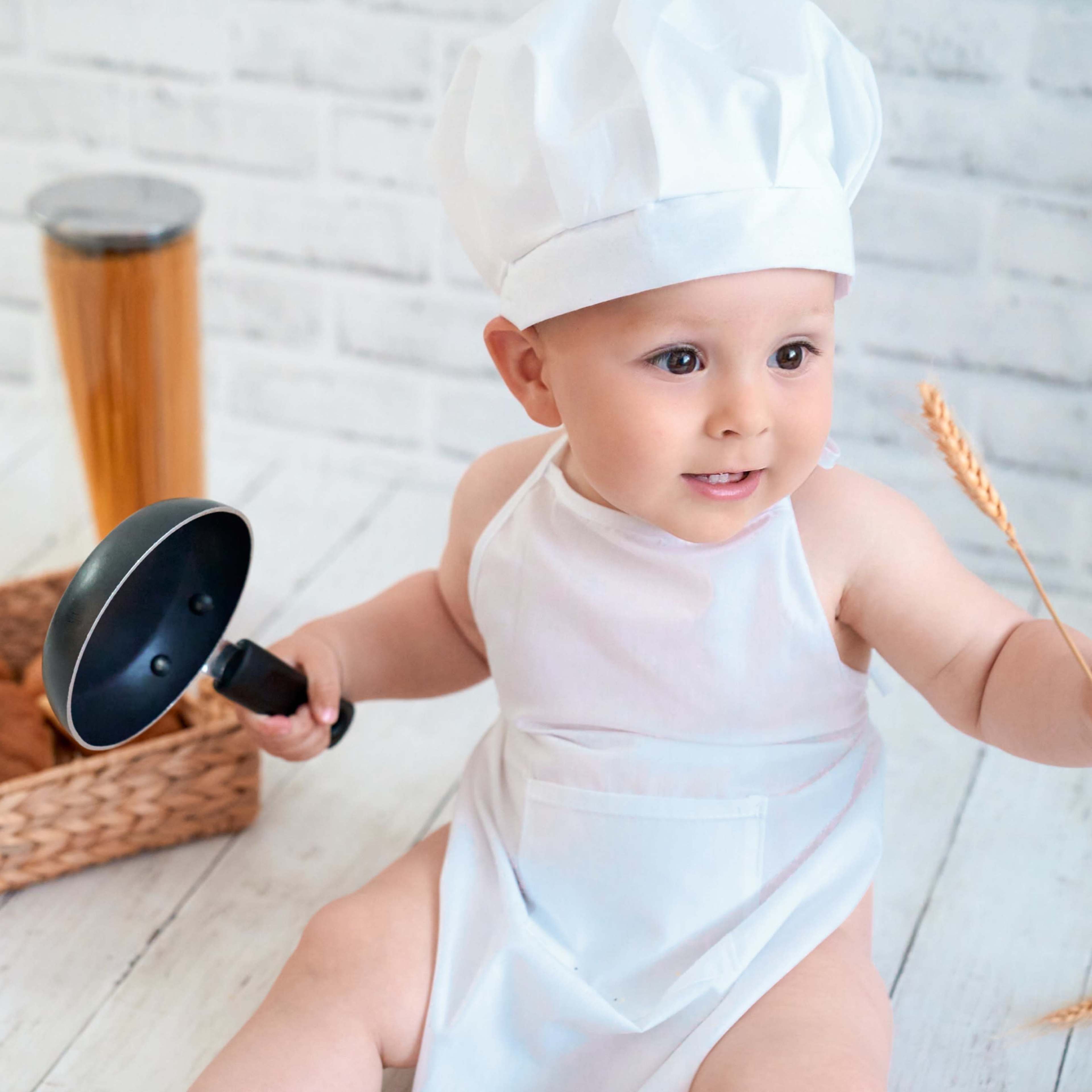 A baby wearing a chef's hat and an apron is sitting on the floor, holding a small frying pan in one hand and a piece of wheat in the other, with a basket of cookies nearby.