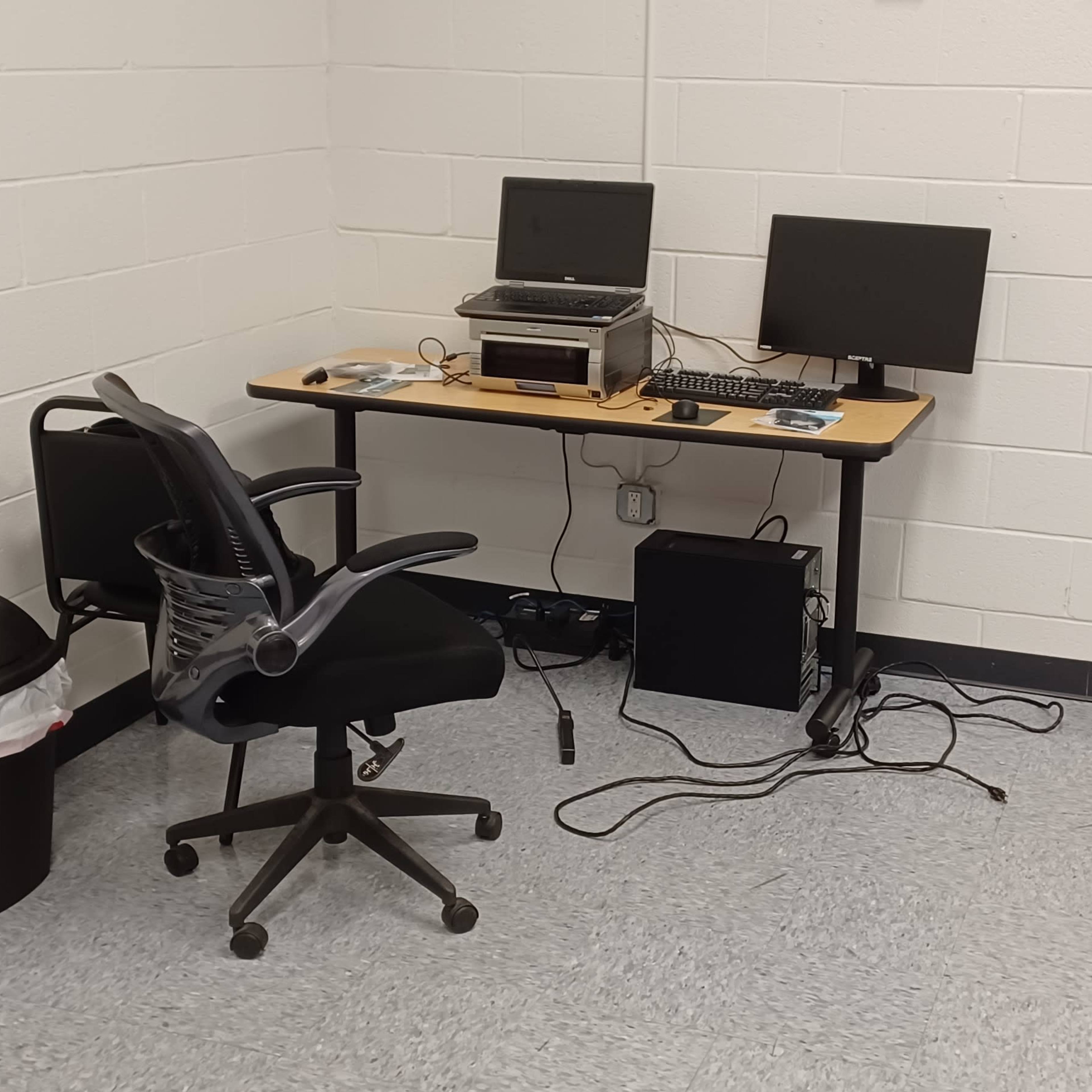 A computer workstation is set up in a corner of a room, featuring two monitors, a printer, an office chair, and a trash bin nearby.