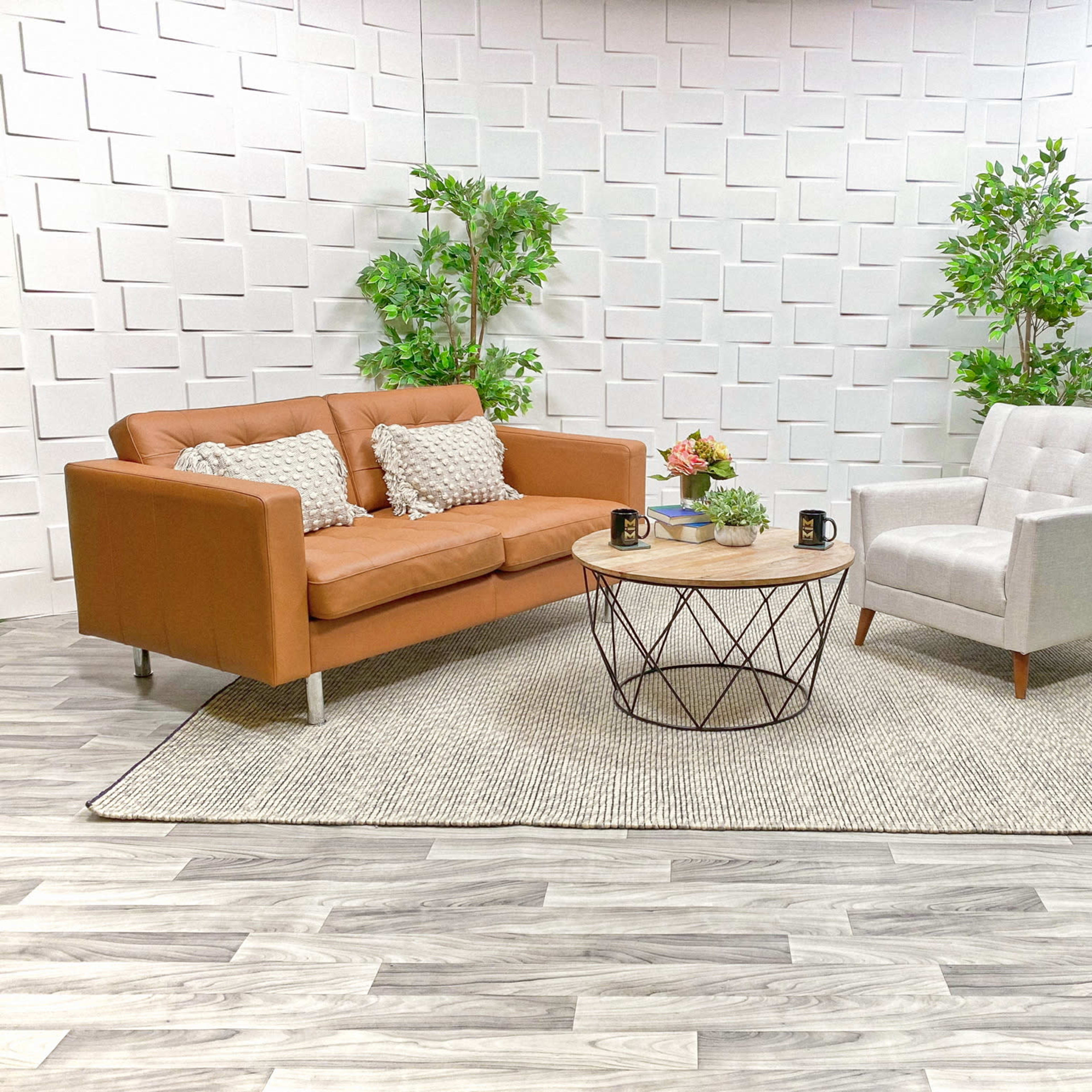 A brown leather sofa and a light gray armchair are arranged around a circular wooden coffee table on a textured rug in a modern, minimalist setting with decorative plants and a geometric wall.