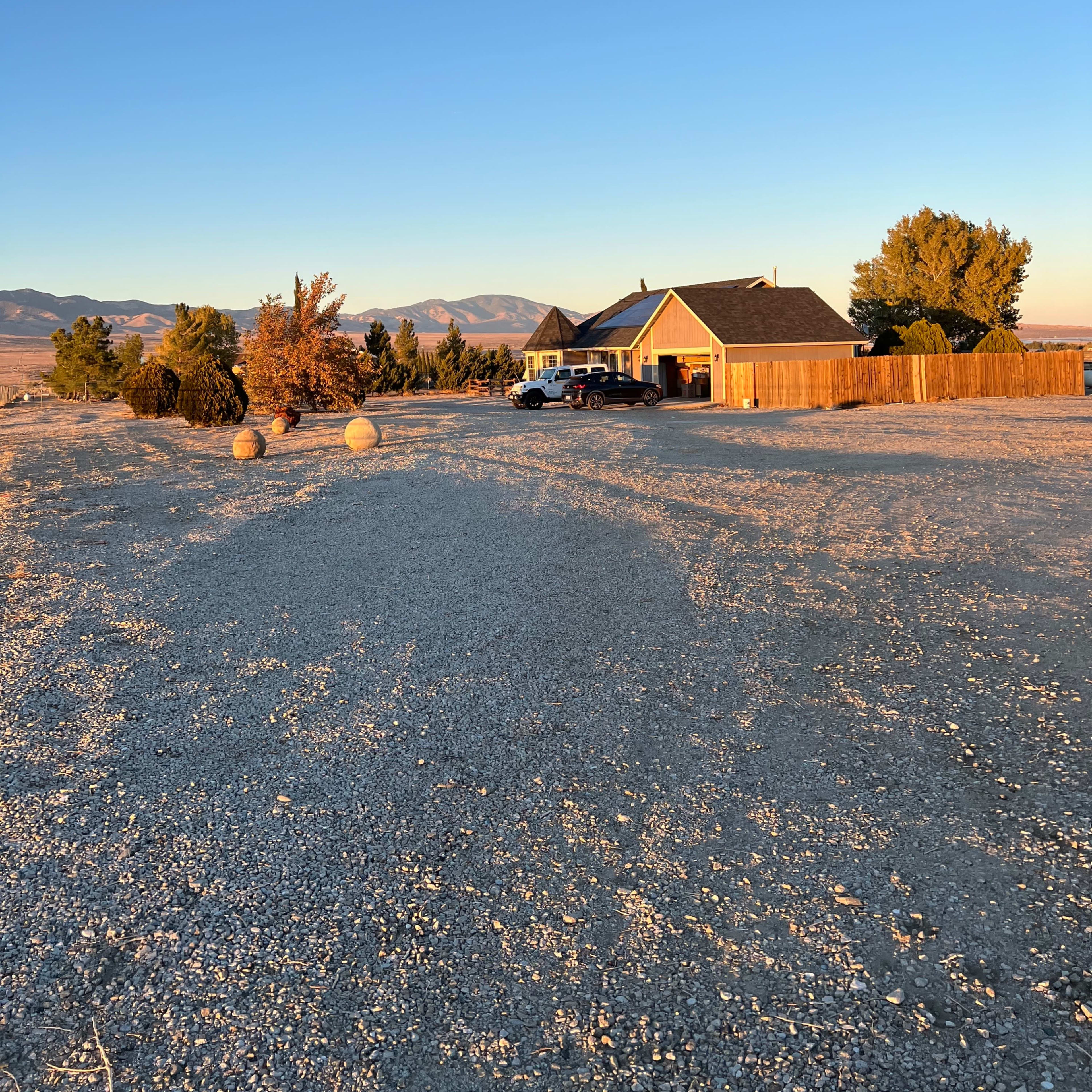 A single-story house is situated on a gravel lot surrounded by trees and distant mountains under a clear blue sky.