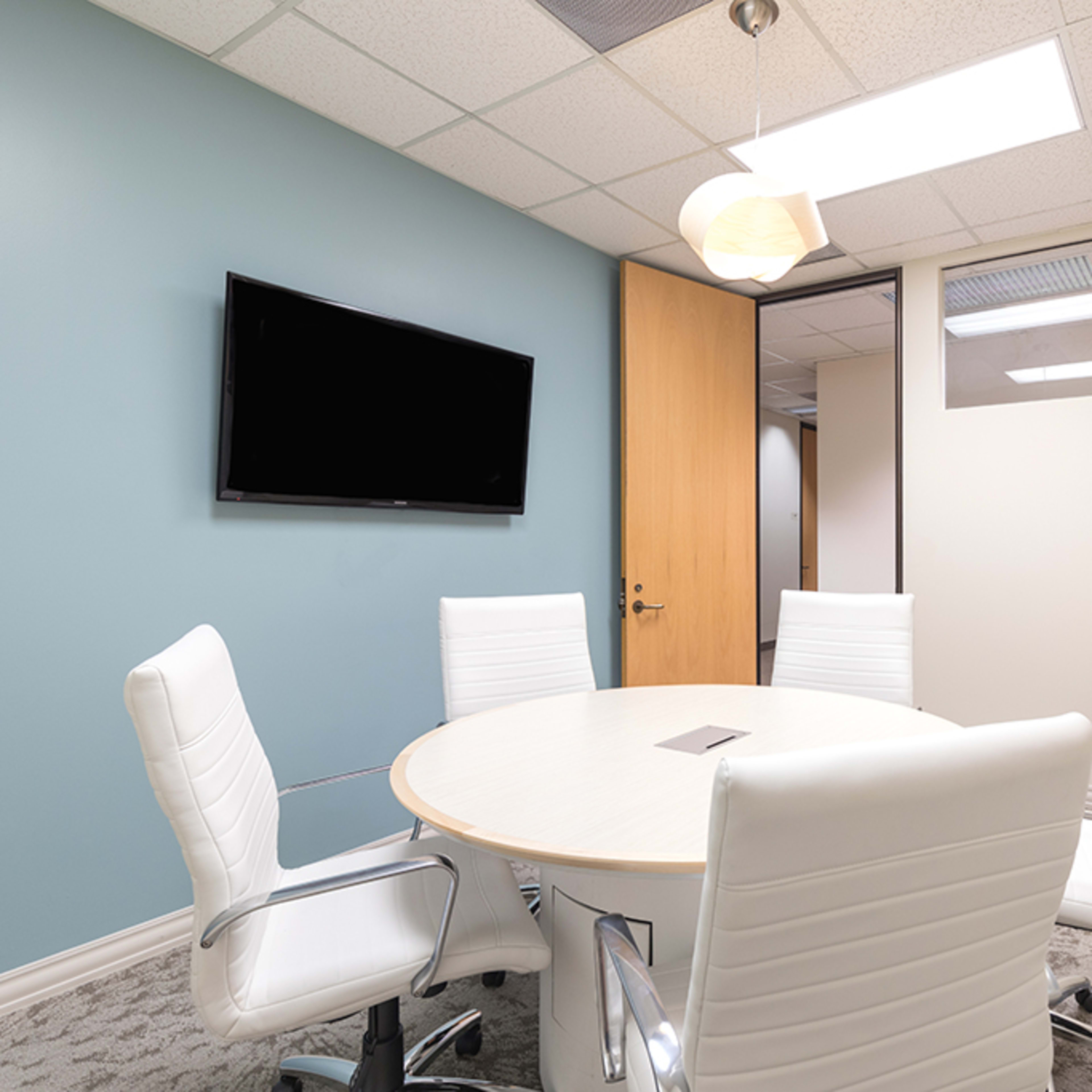 A modern conference room with a round table surrounded by four white chairs, a wall-mounted television, and large windows.