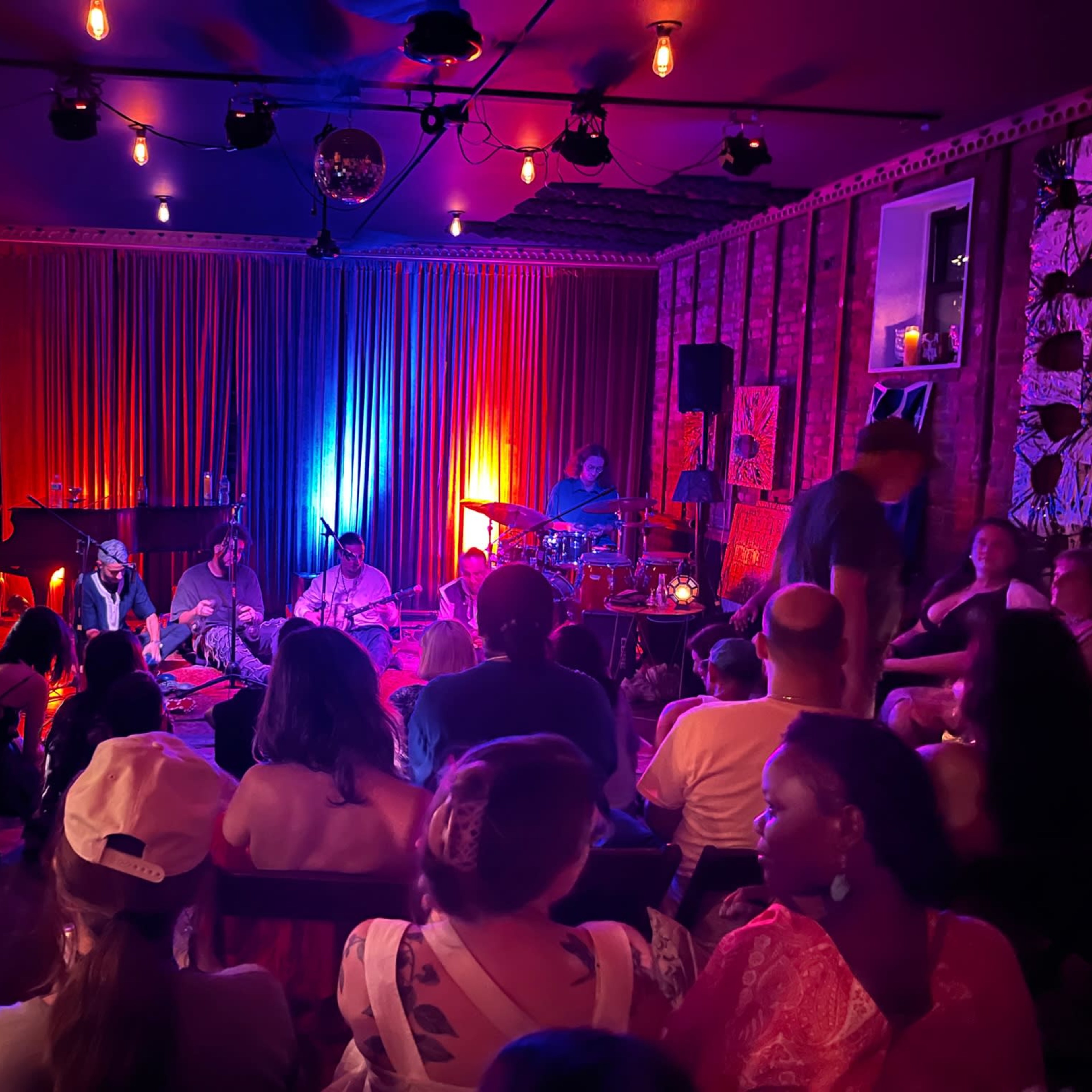 A live music performance in an intimate venue, with musicians playing instruments on stage and an audience seated closely around them.