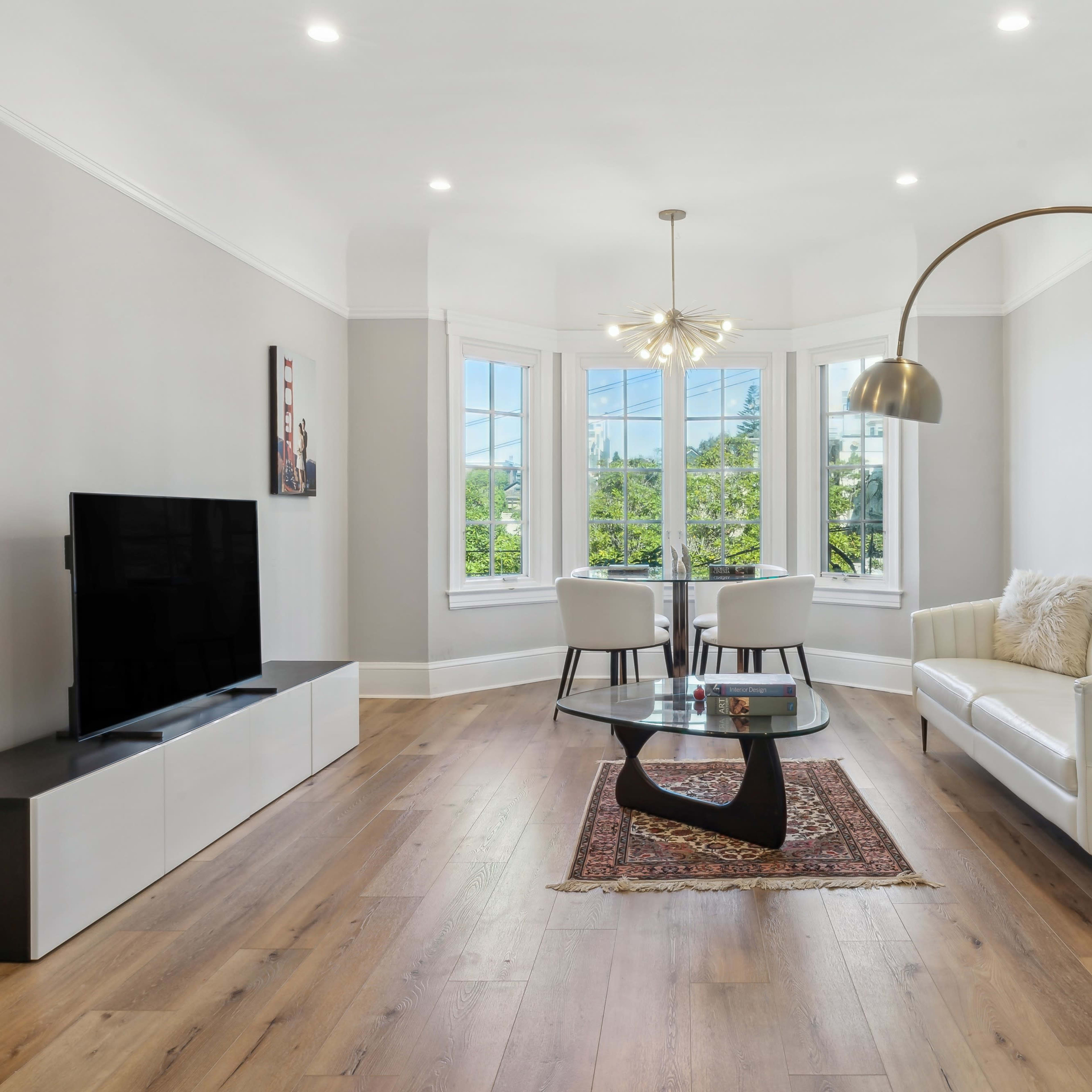 A modern living room features a white sofa, a glass coffee table, a television, and two chairs near large windows.