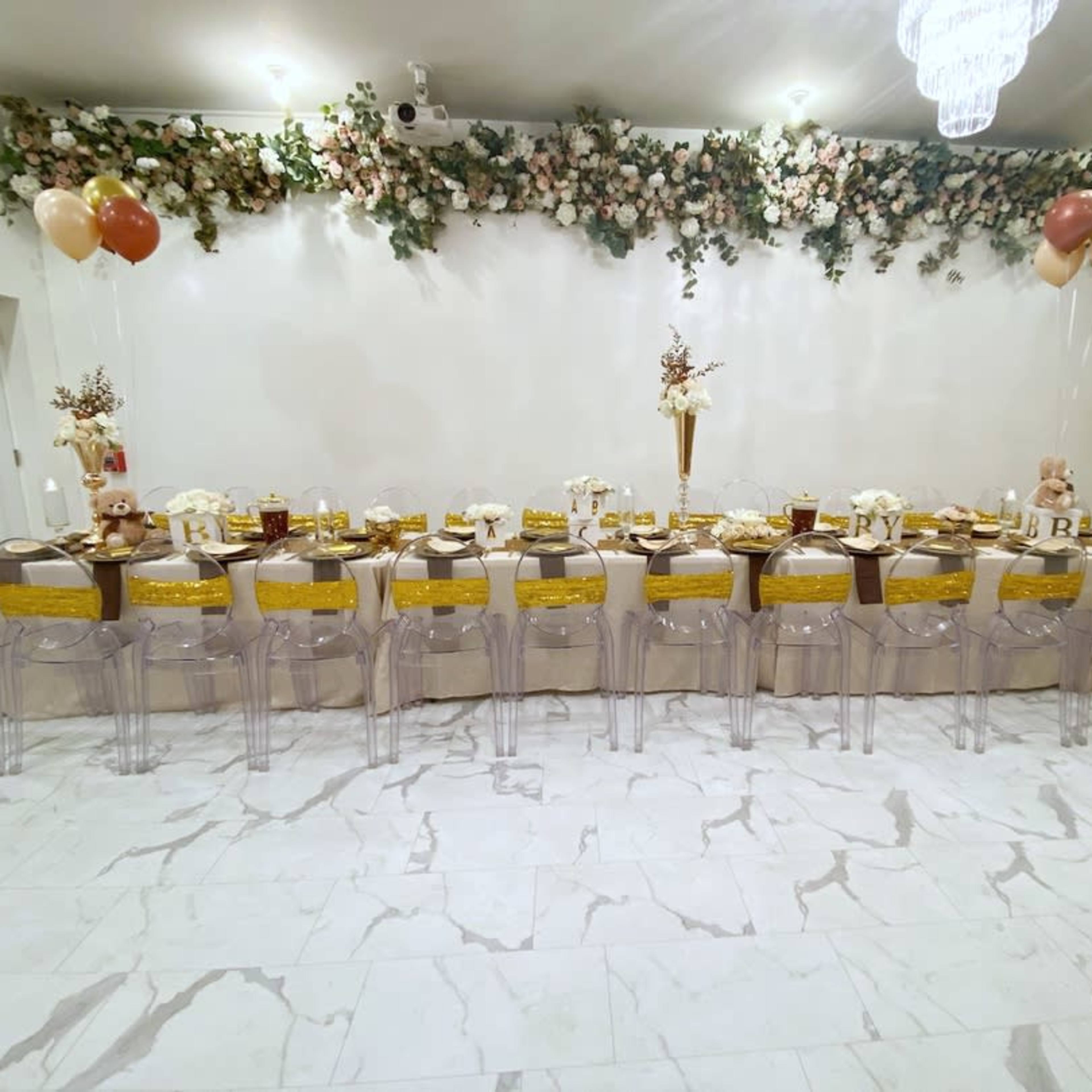 A long banquet table is set with gold and white tableware, surrounded by clear chairs, and decorated with floral arrangements and balloons.