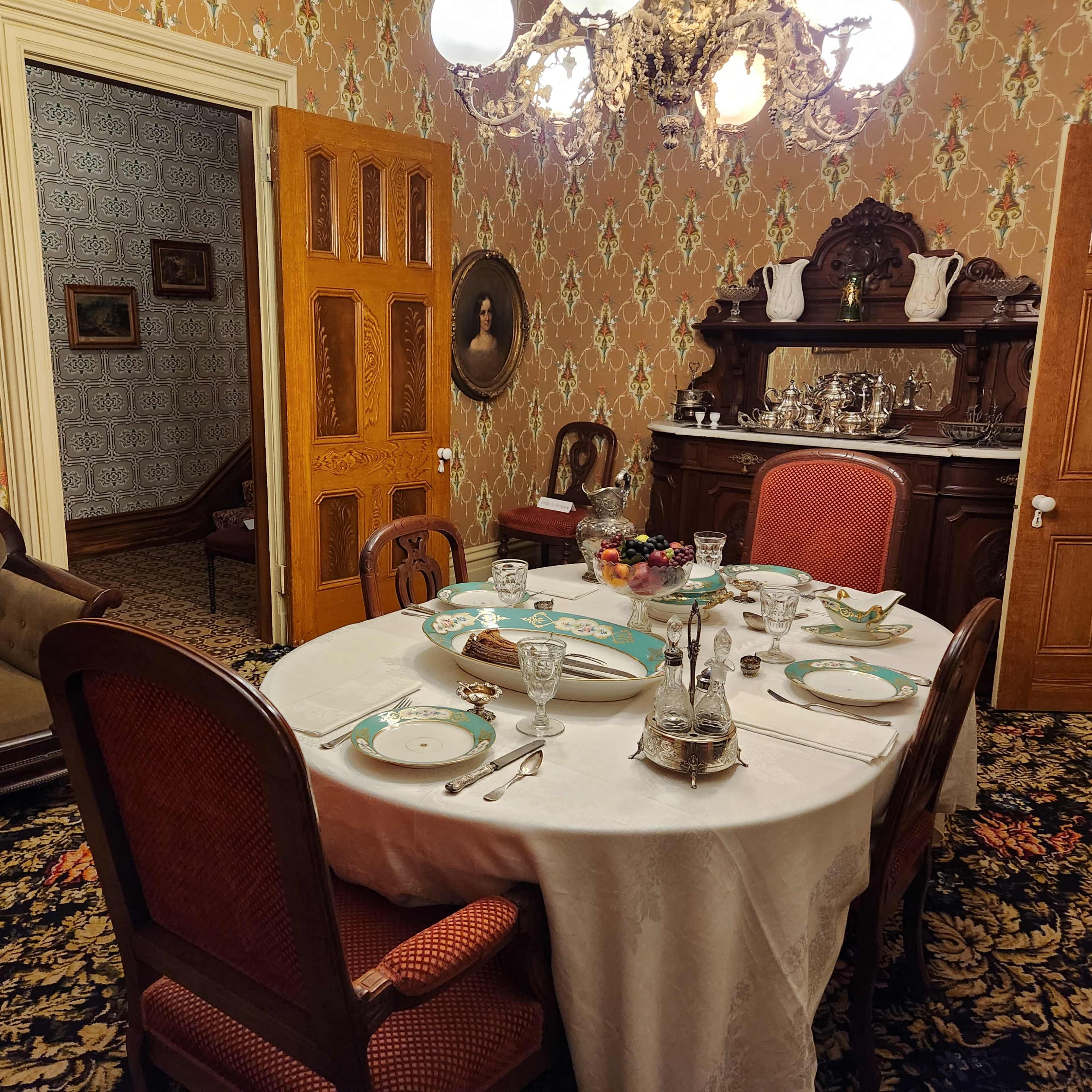 A formal dining room set with a table featuring an ornate tablecloth, china, and glassware, surrounded by wooden chairs against a backdrop of patterned wallpaper and vintage decor.