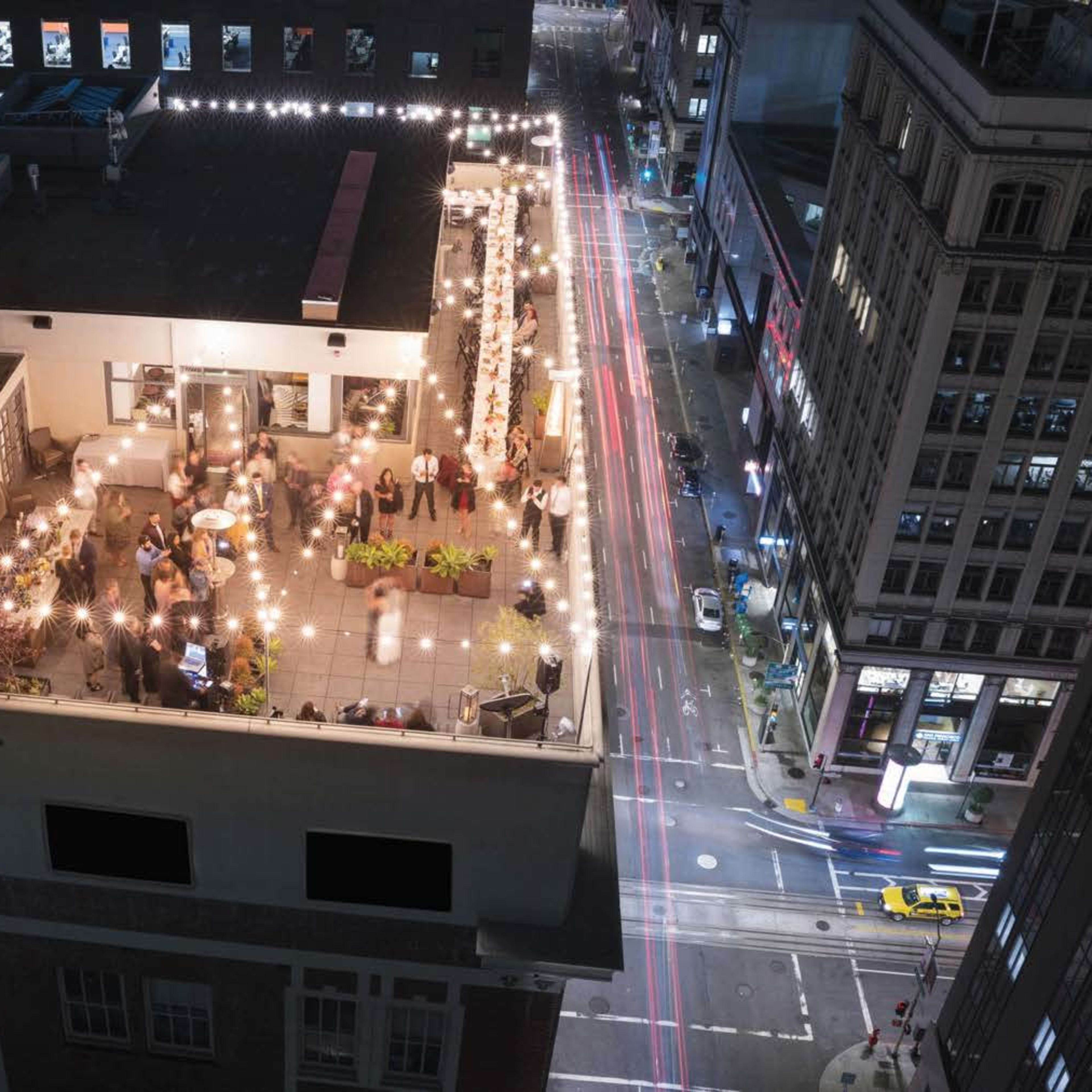 A rooftop bar filled with patrons and string lights overlooks a busy city street at night.