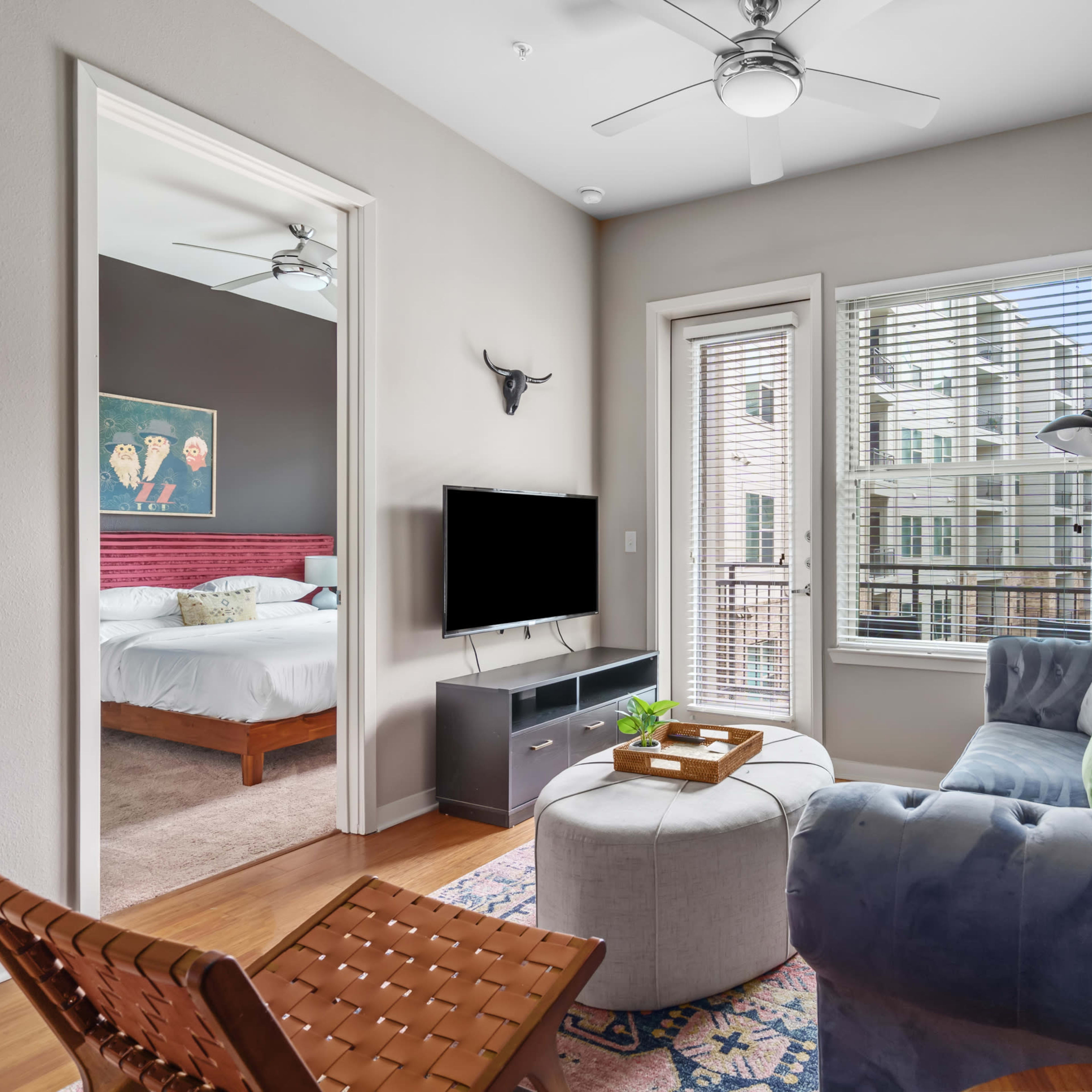 A living room features a blue sofa, a round ottoman, a television on a stand, and an adjoining bedroom visible through a set of double doors.