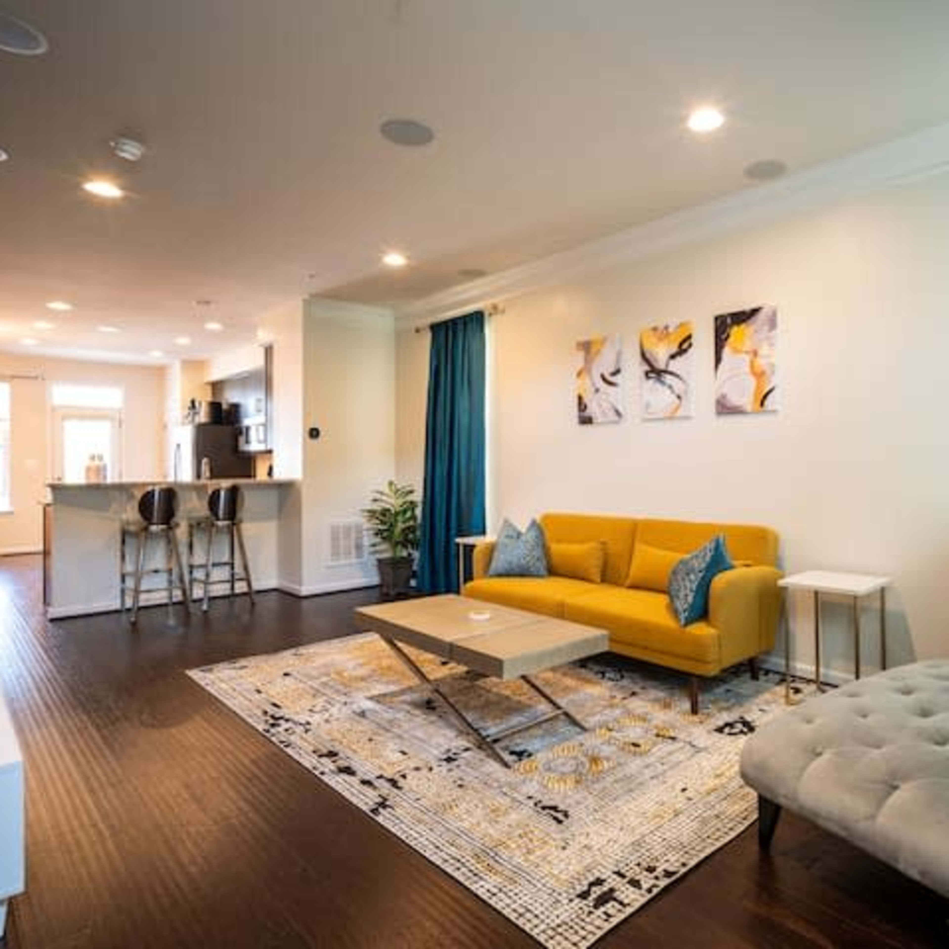 A modern living room features a yellow sofa, gray chaise lounge, and a patterned area rug, with a kitchen bar visible in the background.