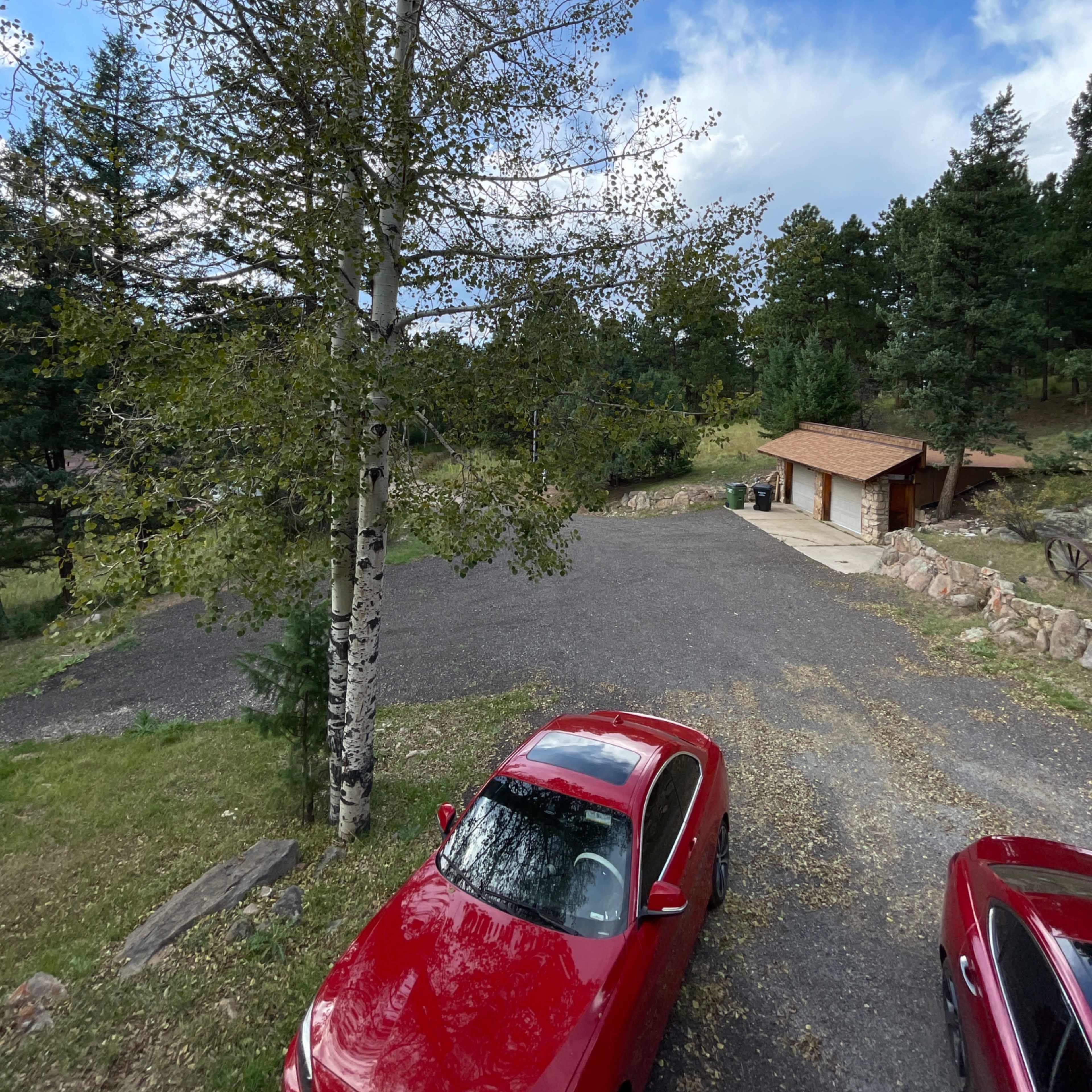 A view of a gravel parking area with two red cars parked in the foreground, bordered by trees and a small building in the background.
