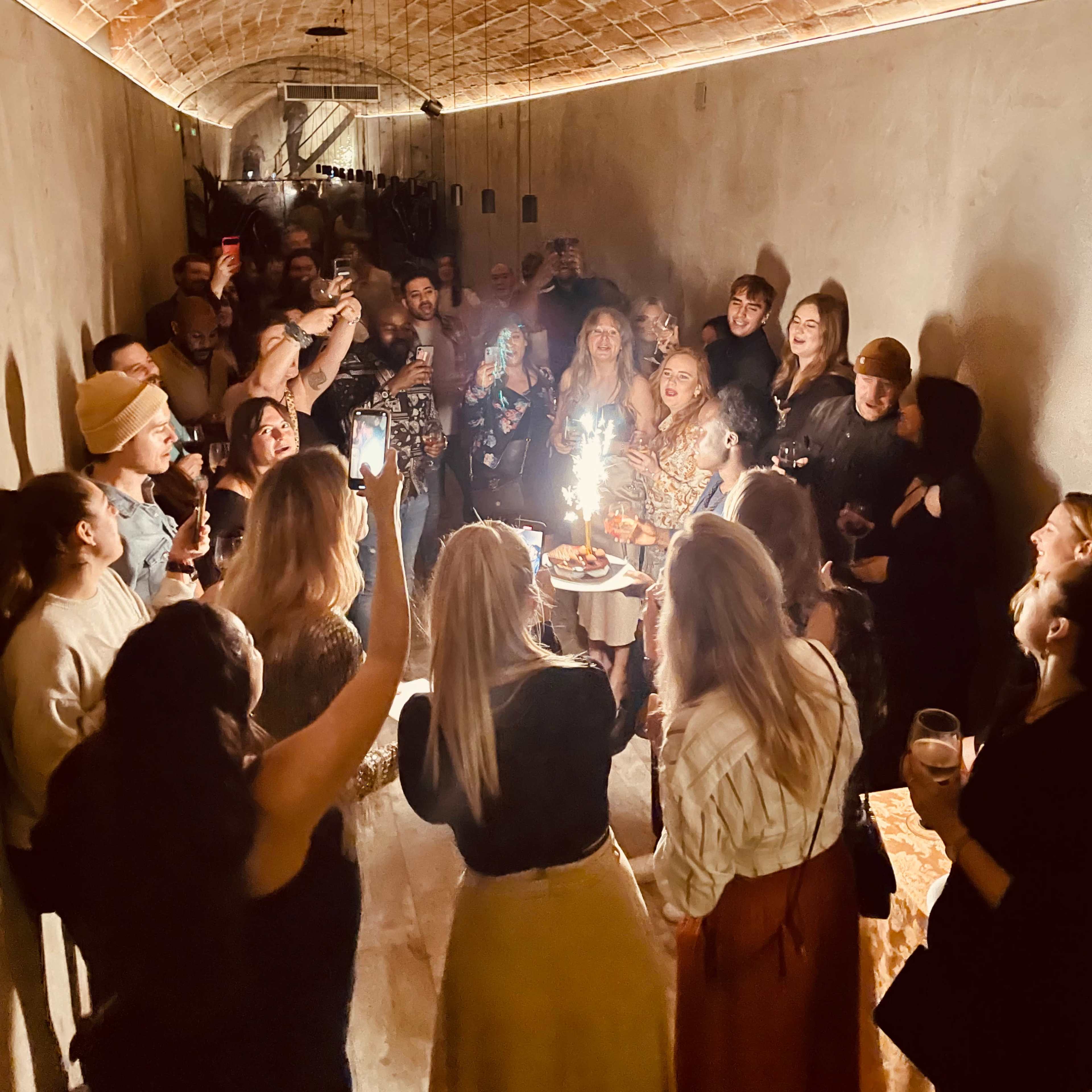 A crowd of people gathers in a dimly lit arched hallway, celebrating with drinks and a birthday cake topped with candles.