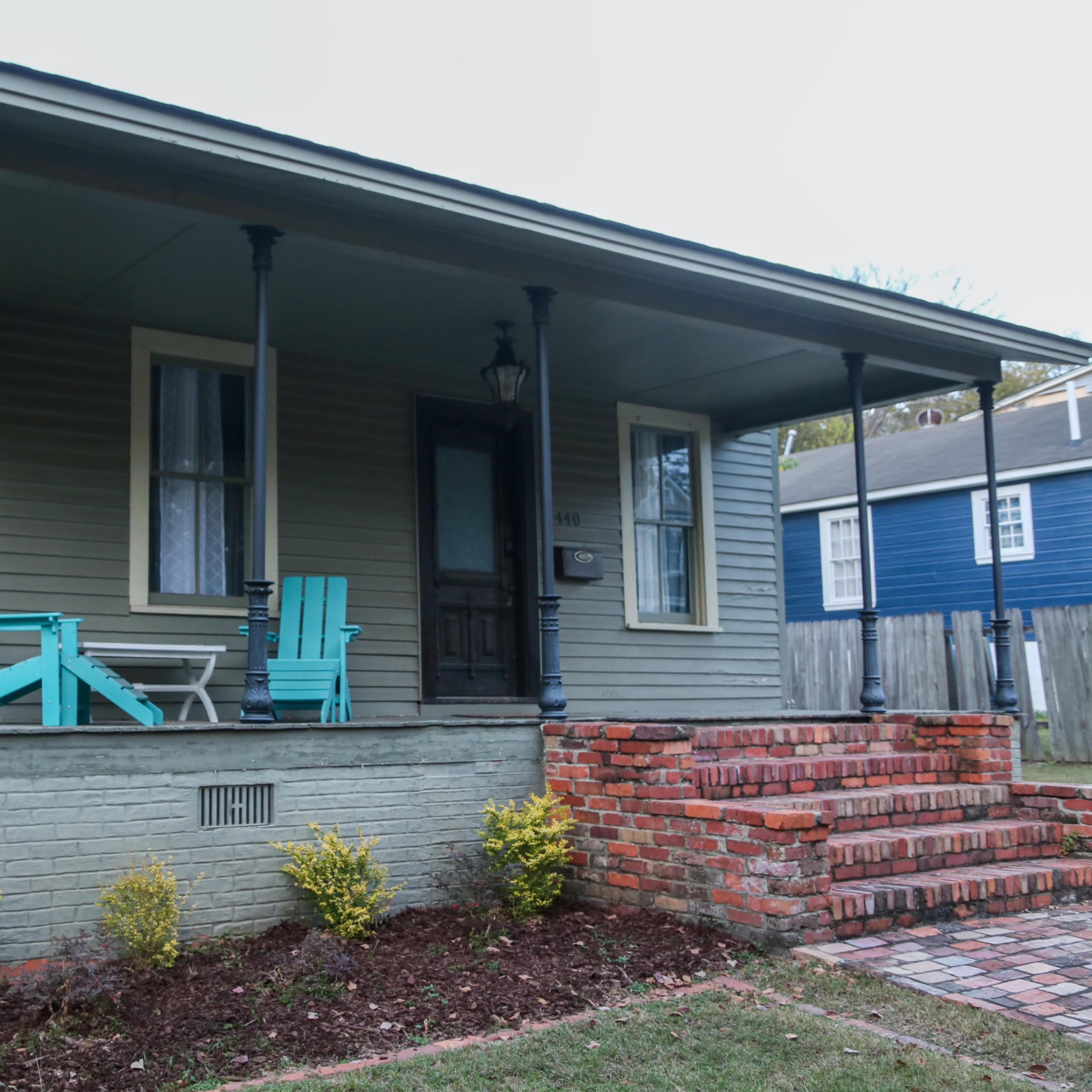 A light green house with a front porch featuring two turquoise chairs and steps made of red bricks leading to the entrance.