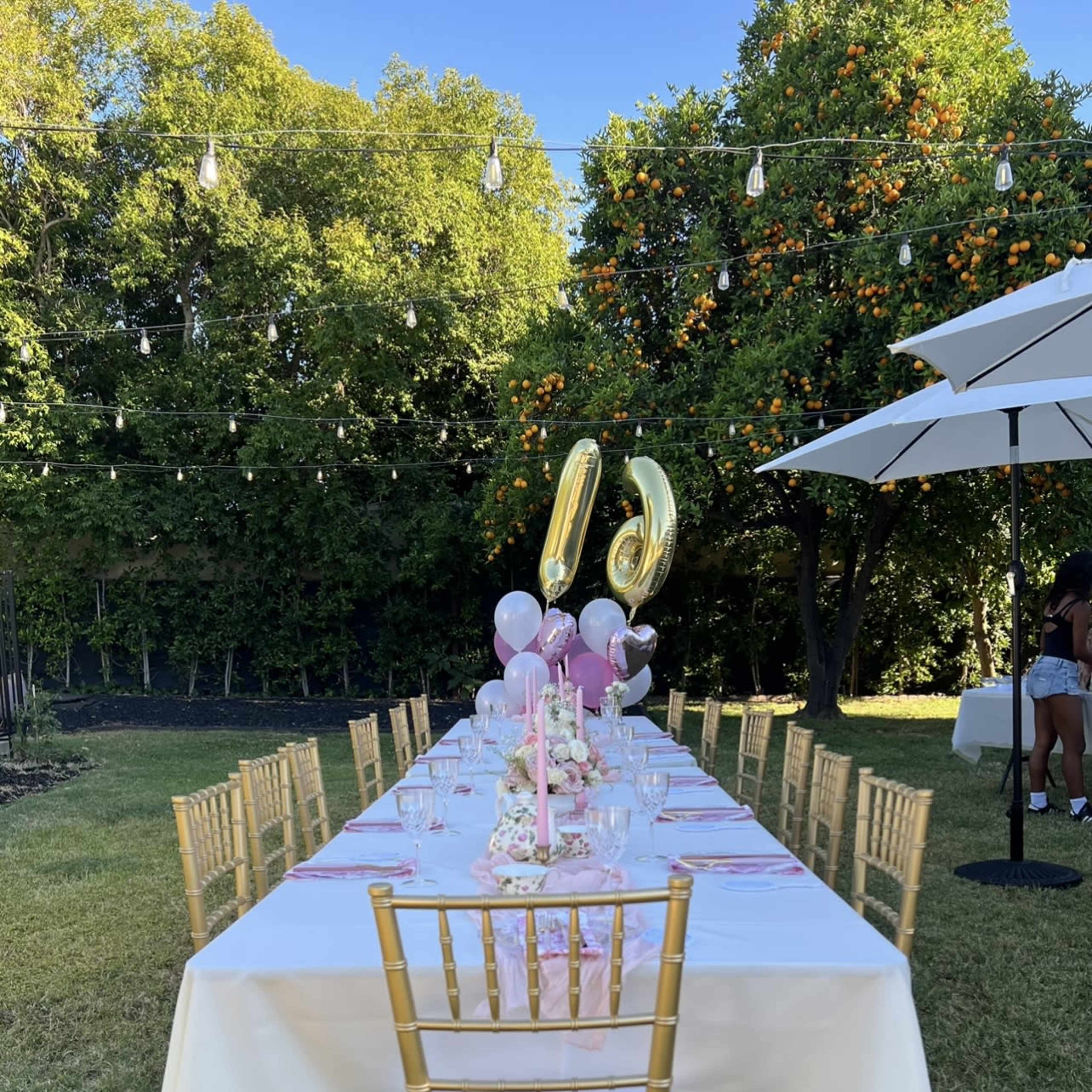 A long dining table is set outdoors with pink decorations, golden balloons, and chairs under string lights and orange trees.