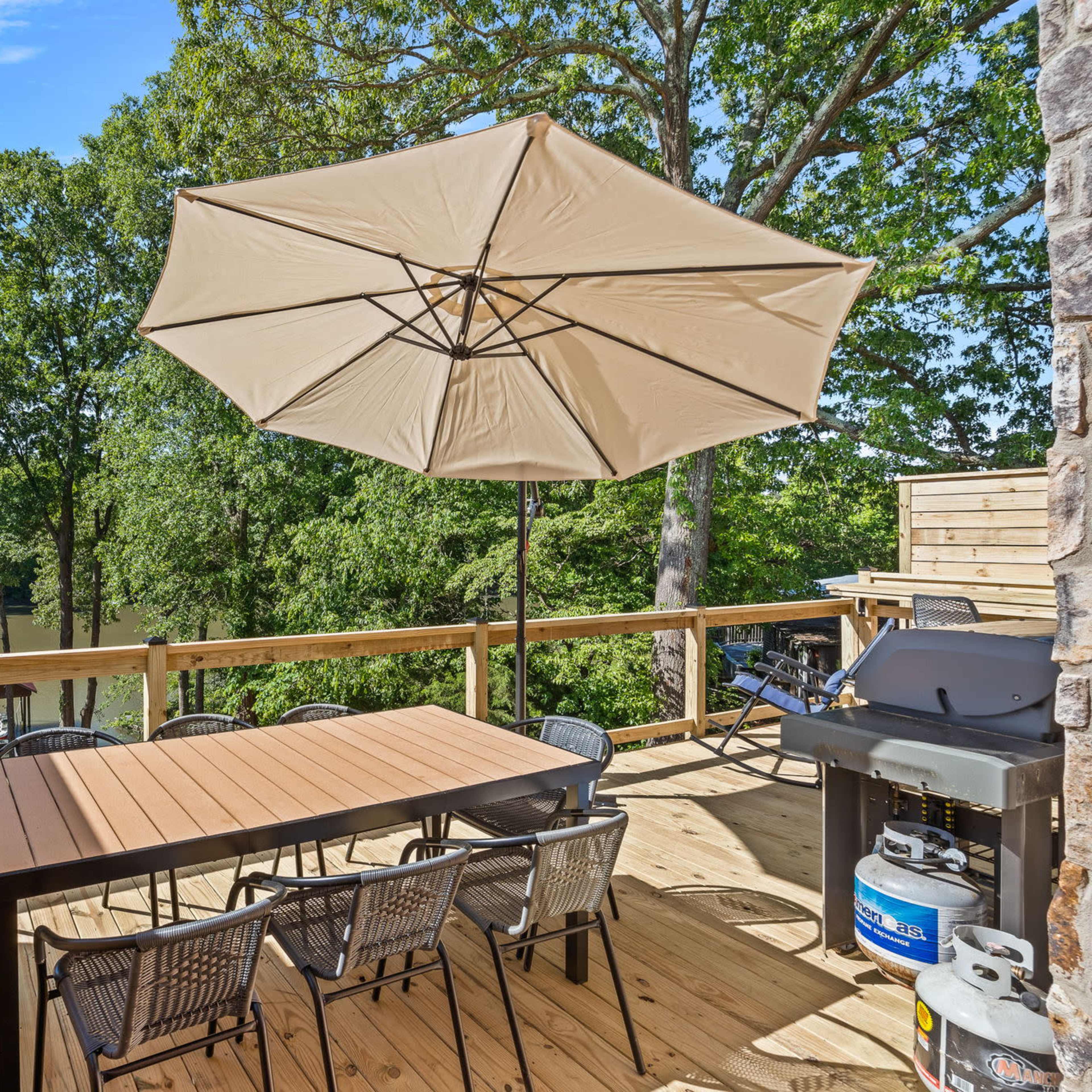 A wooden deck with a table and chairs is shaded by a large umbrella, overlooking a calm body of water surrounded by trees.