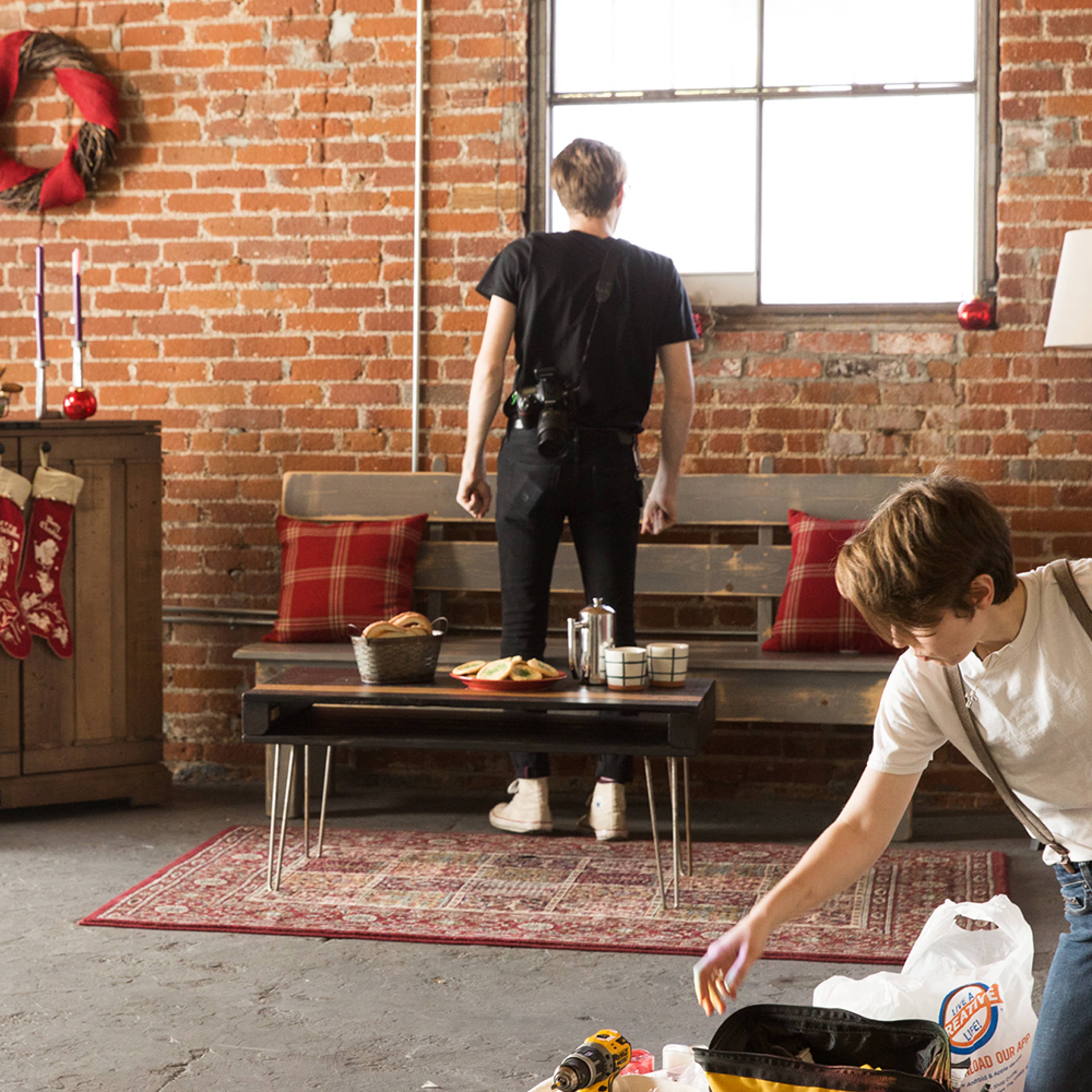A camera setup is positioned in a rustic room where one person stands by a window while another is unpacking a bag on the floor.