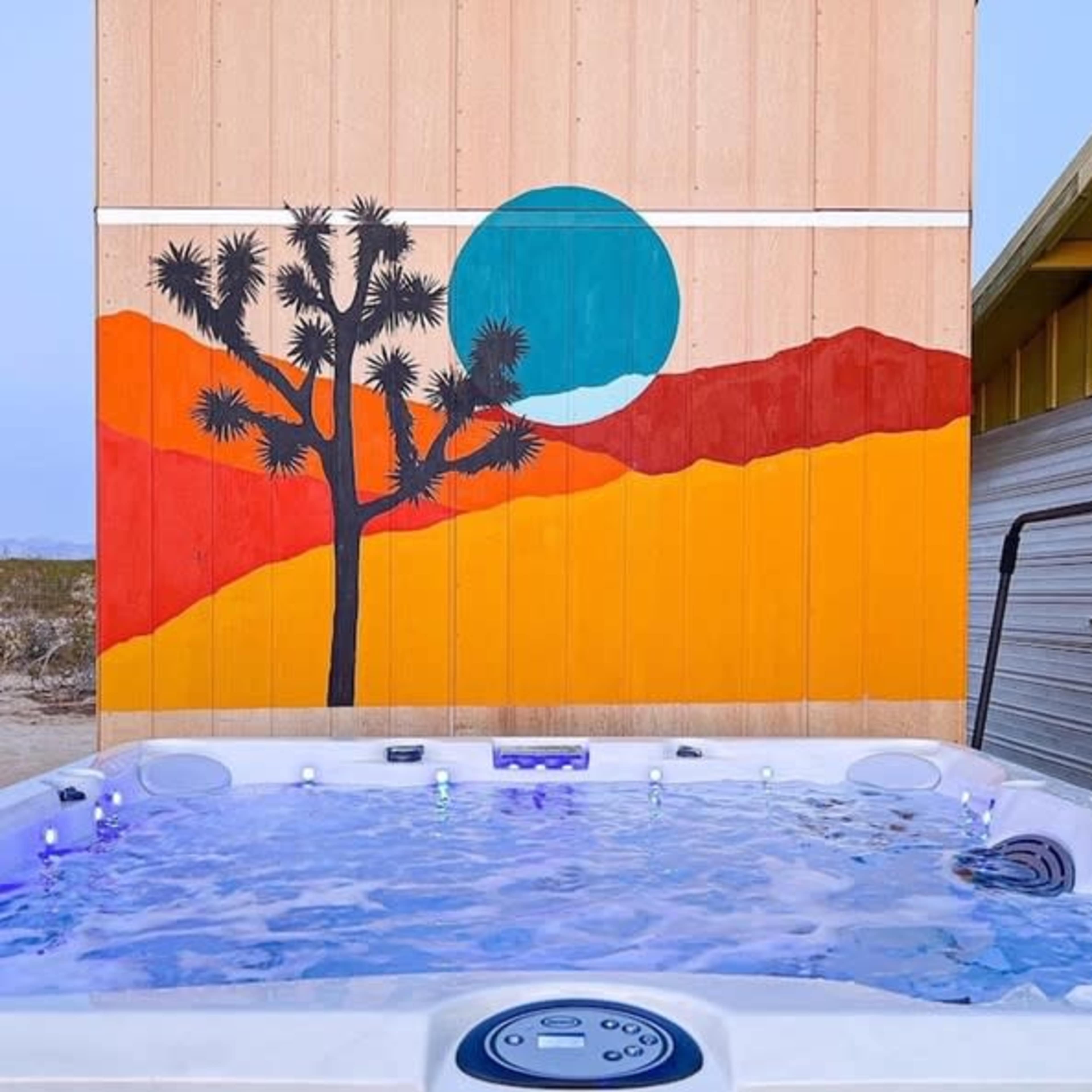 A hot tub in front of a colorful mural depicting a tree and a sunset in a desert landscape.