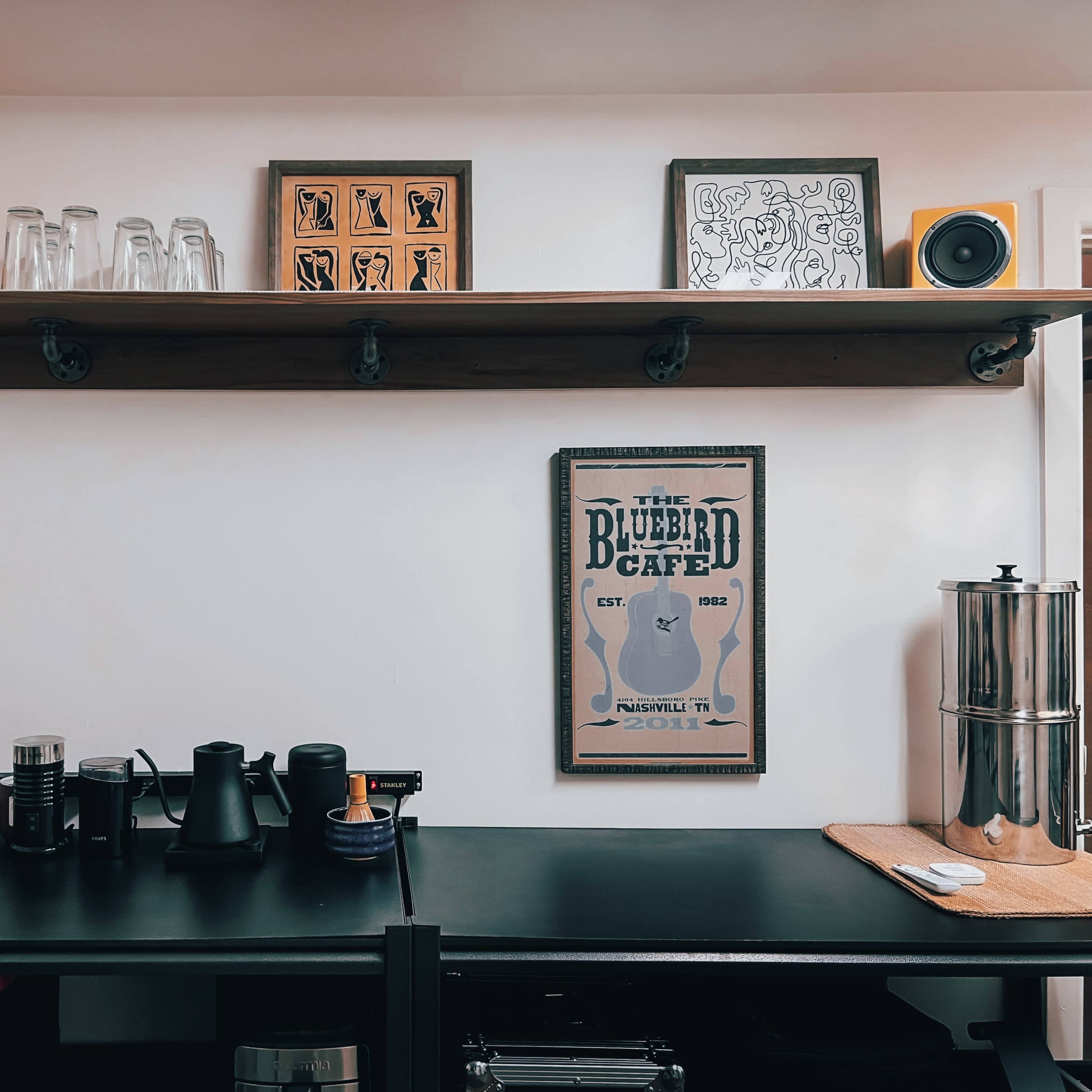 A kitchen counter with various bottles, coffee equipment, and a shelf above displaying glassware and speakers, along with a framed poster.