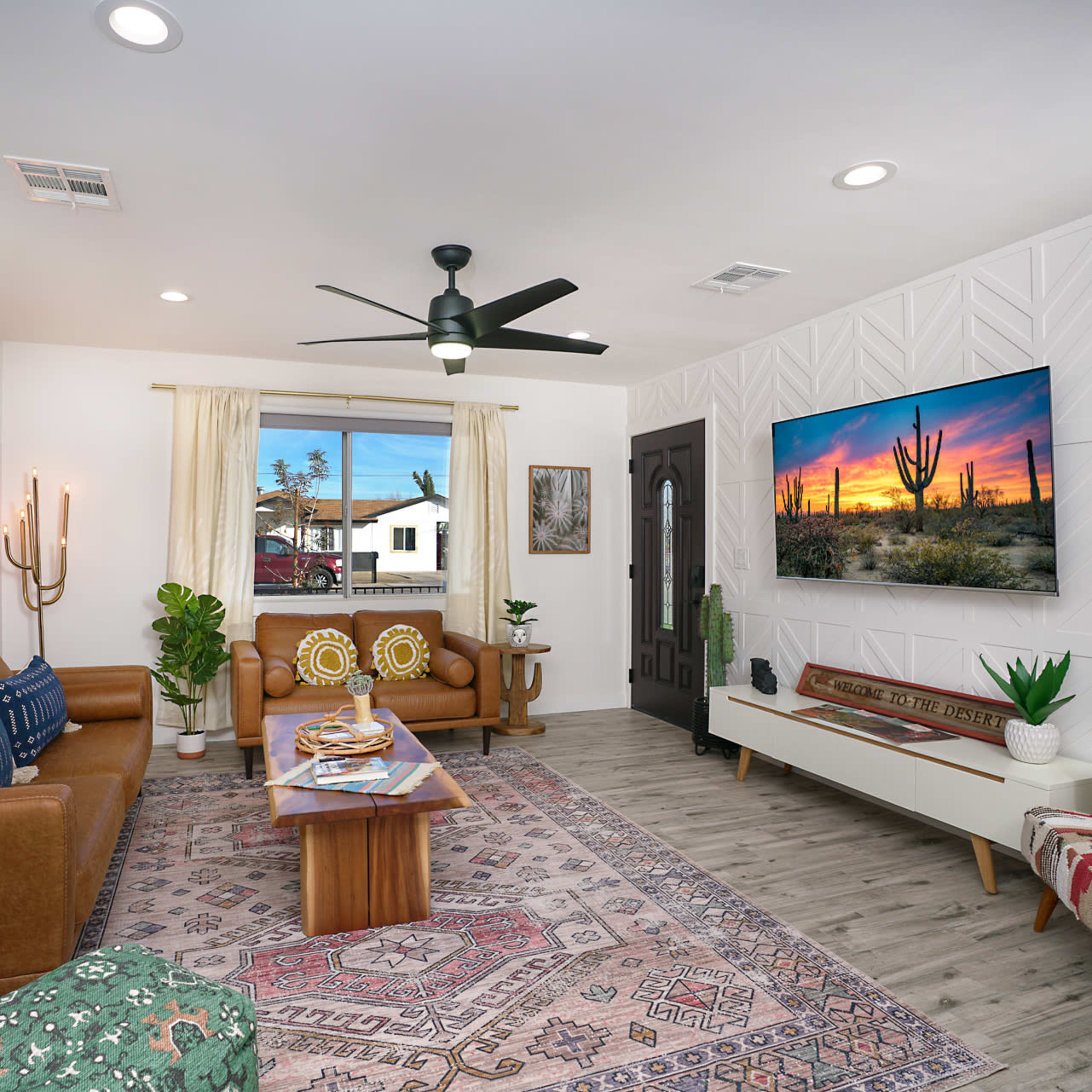 A living room features brown leather sofas, a wooden coffee table, a decorative rug, and a wall-mounted television displaying a desert landscape.