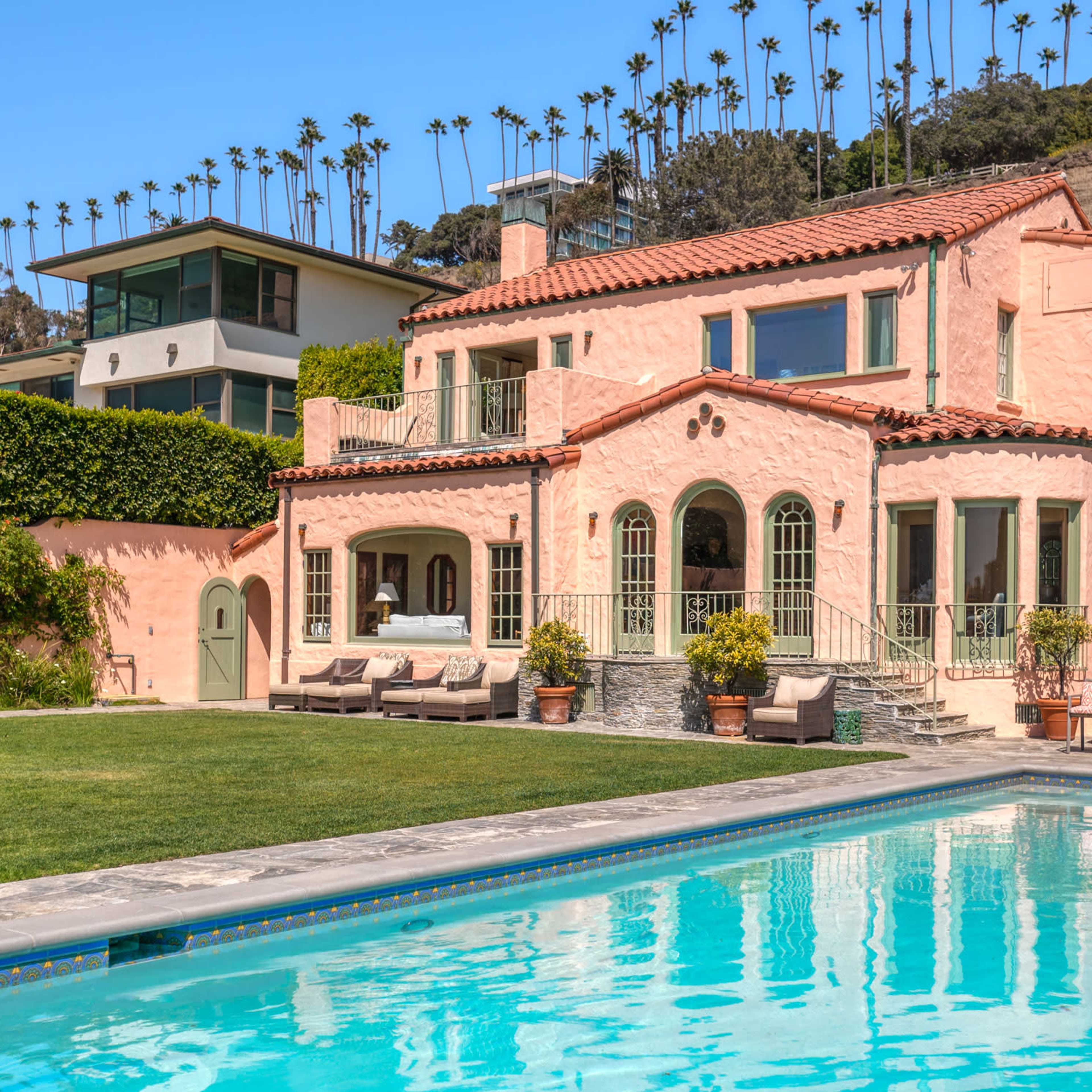 A two-story pink stucco house with arched windows and a pool is surrounded by a manicured lawn and palm trees.
