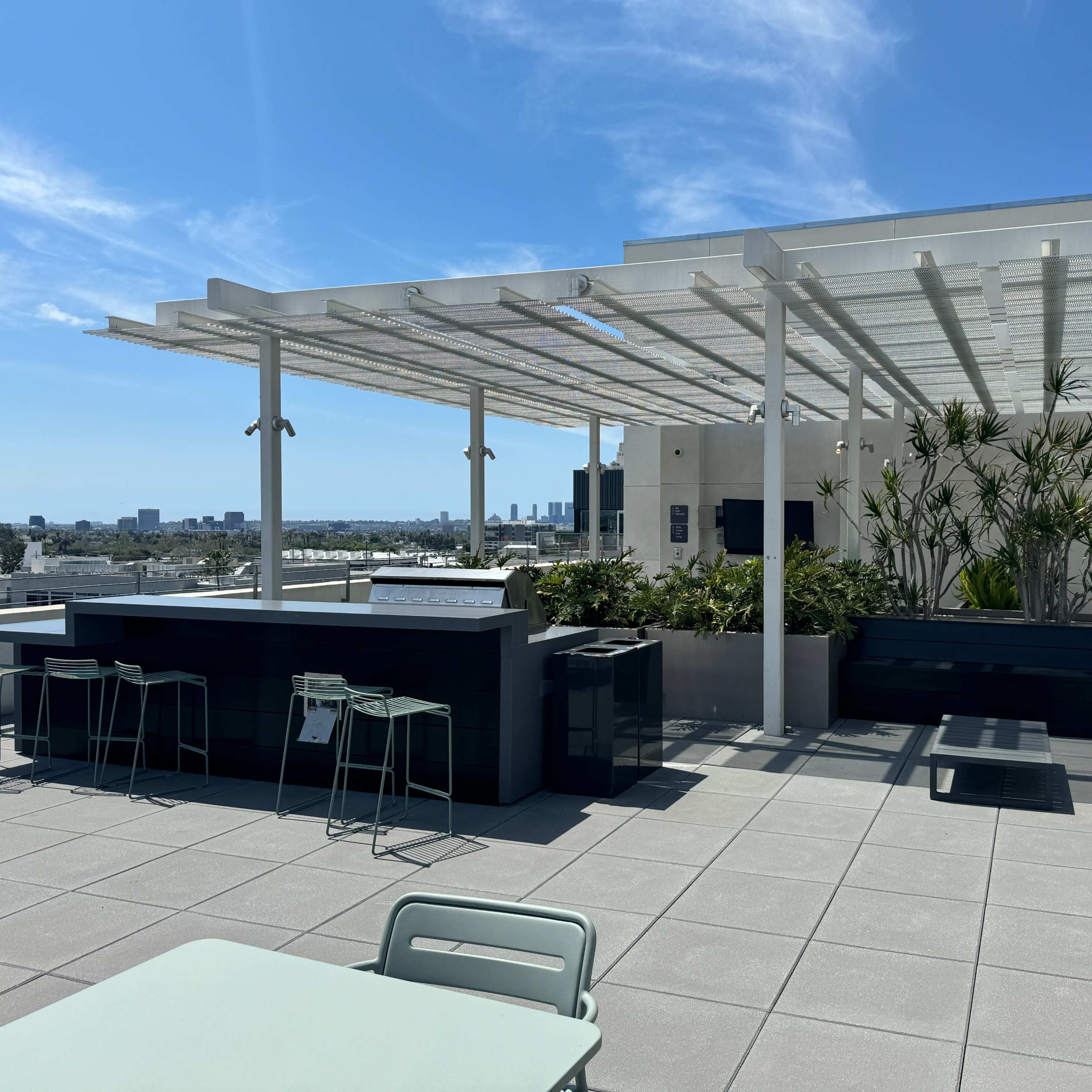 A rooftop terrace featuring a bar area, seating, and potted greenery, with a city skyline visible in the background.