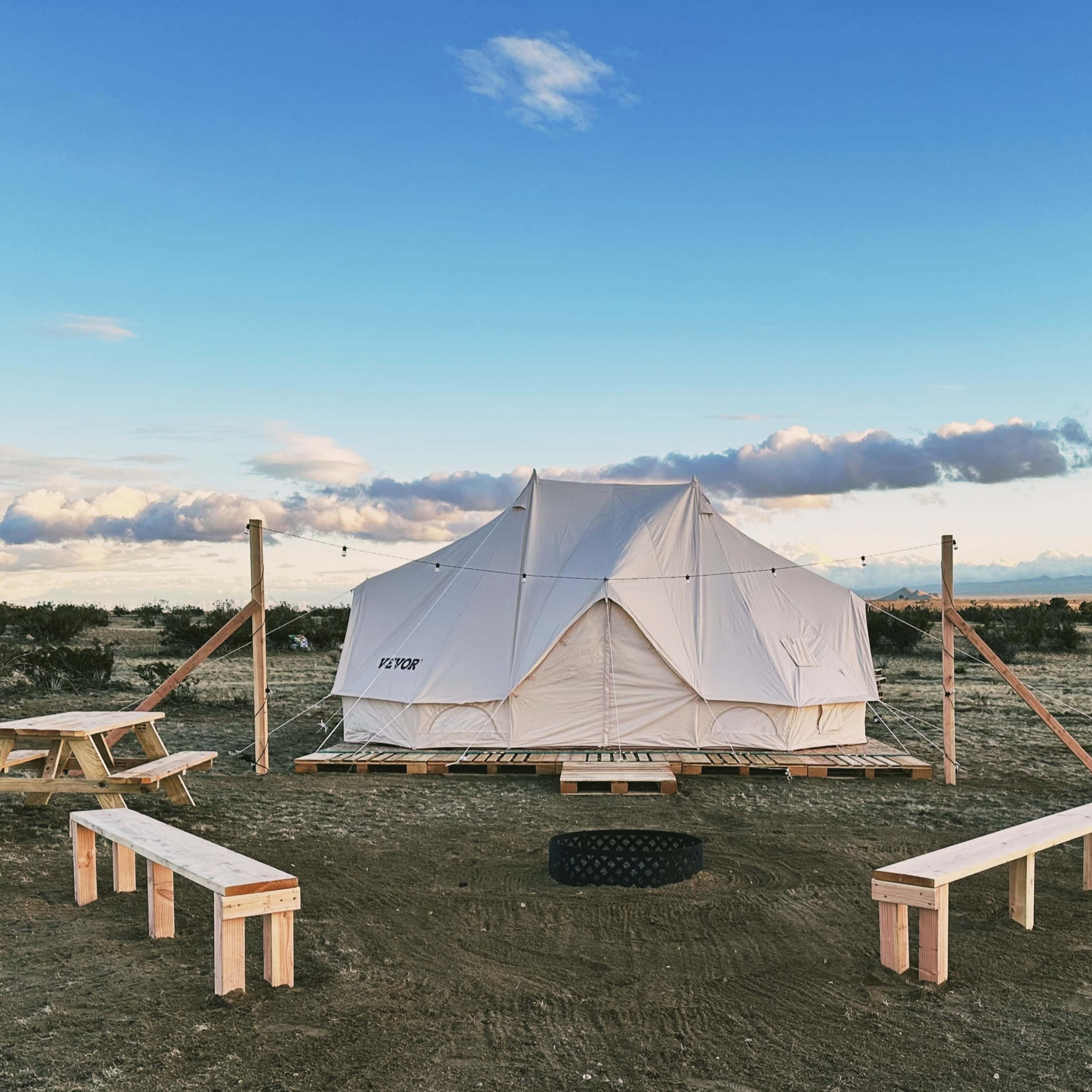 A white tent is set on a wooden platform in a desert landscape, surrounded by picnic tables and a fire pit.