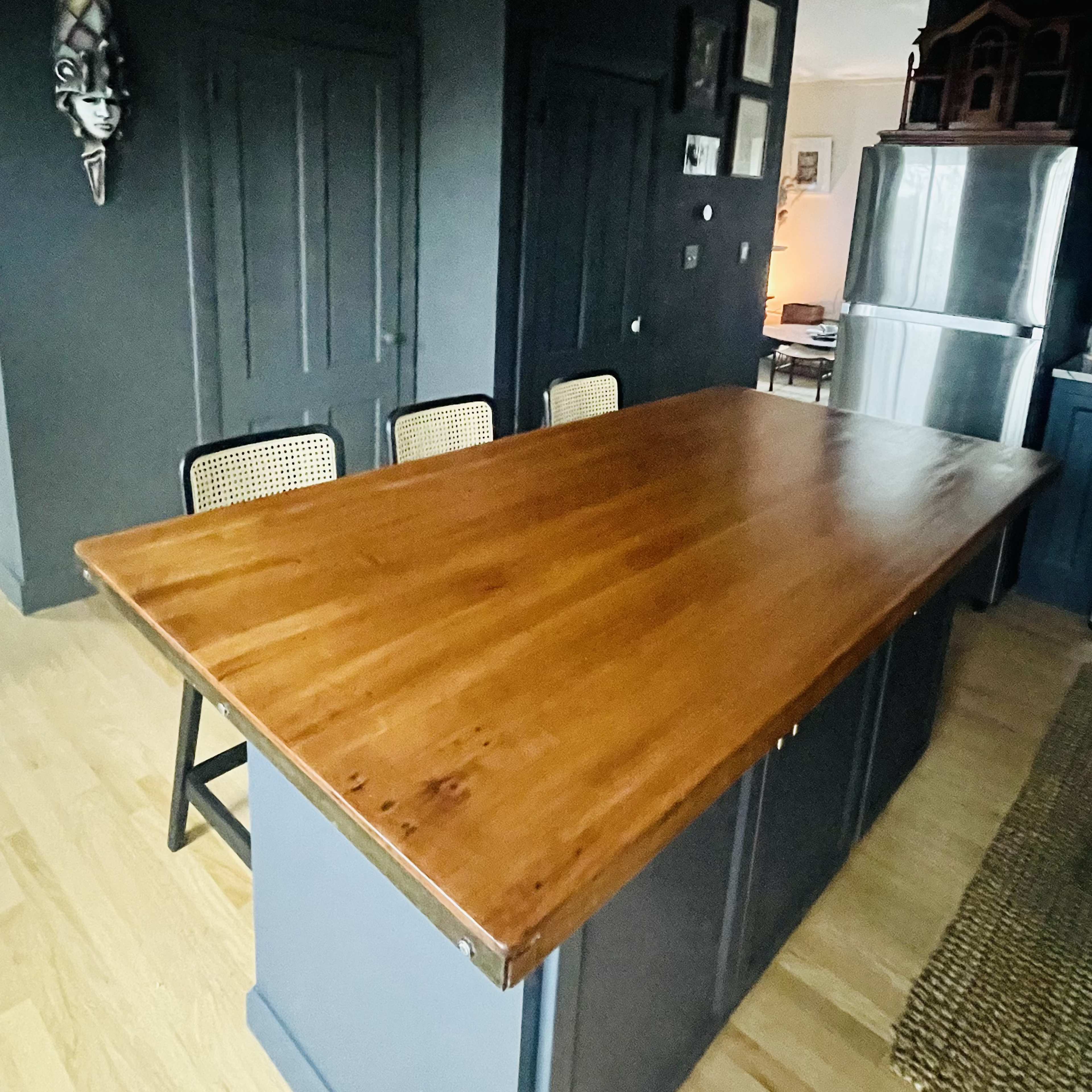 A wooden dining table with a smooth surface is positioned in a dimly lit room, surrounded by cane-back chairs, against dark walls and near a silver refrigerator.