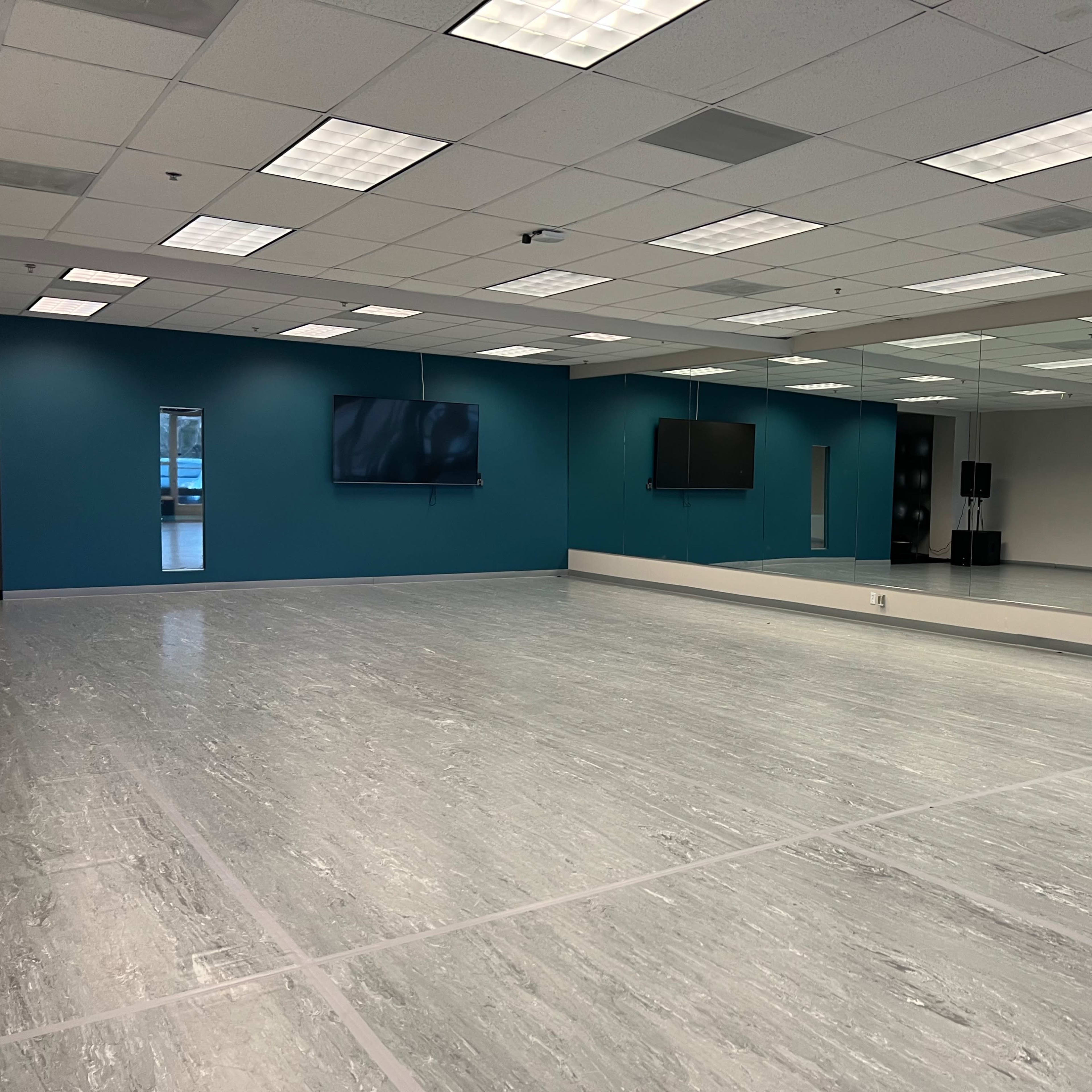 An empty dance studio with large mirrors, two wall-mounted screens, and a light-colored floor.