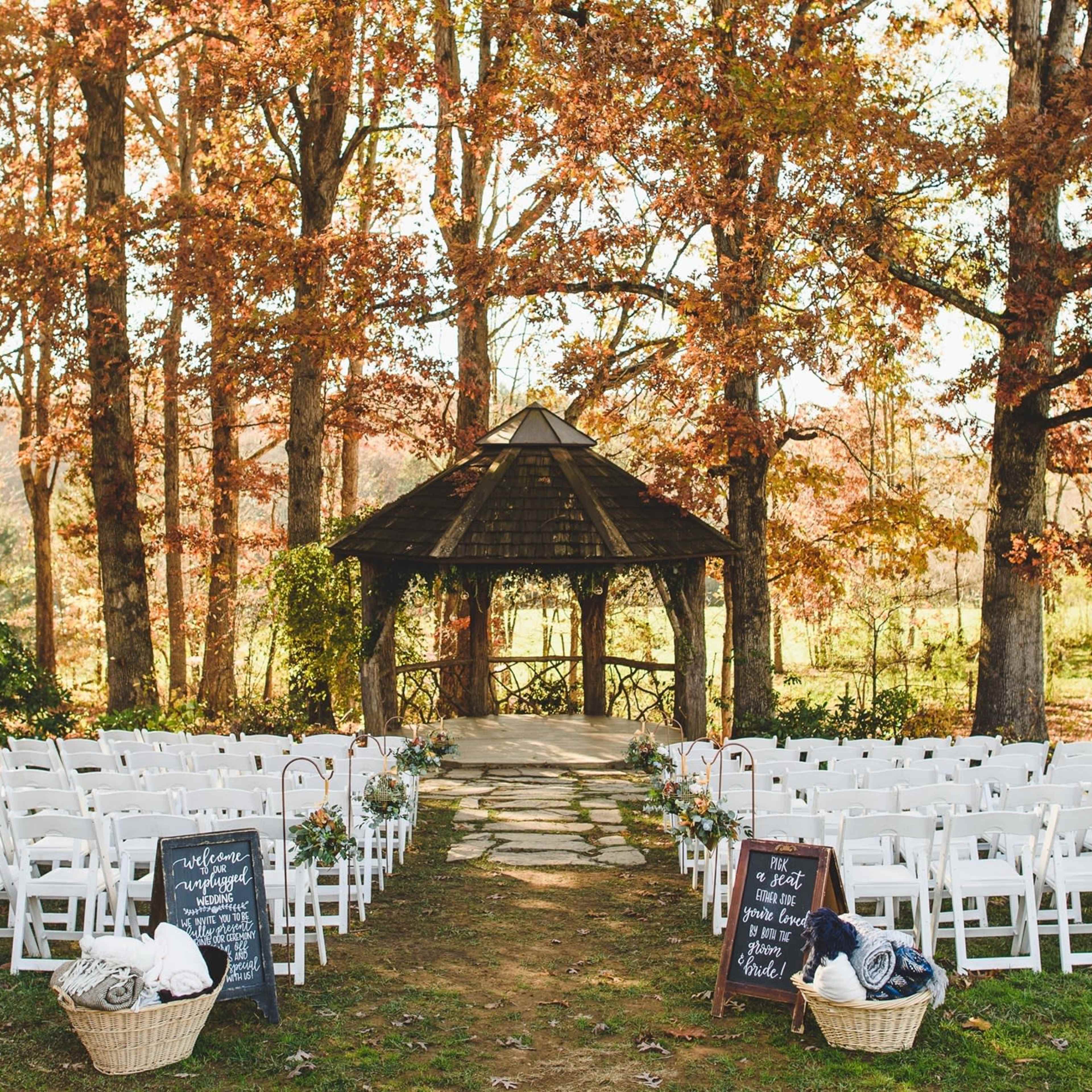 A wedding ceremony setup features rows of white chairs facing a gazebo surrounded by autumn foliage.