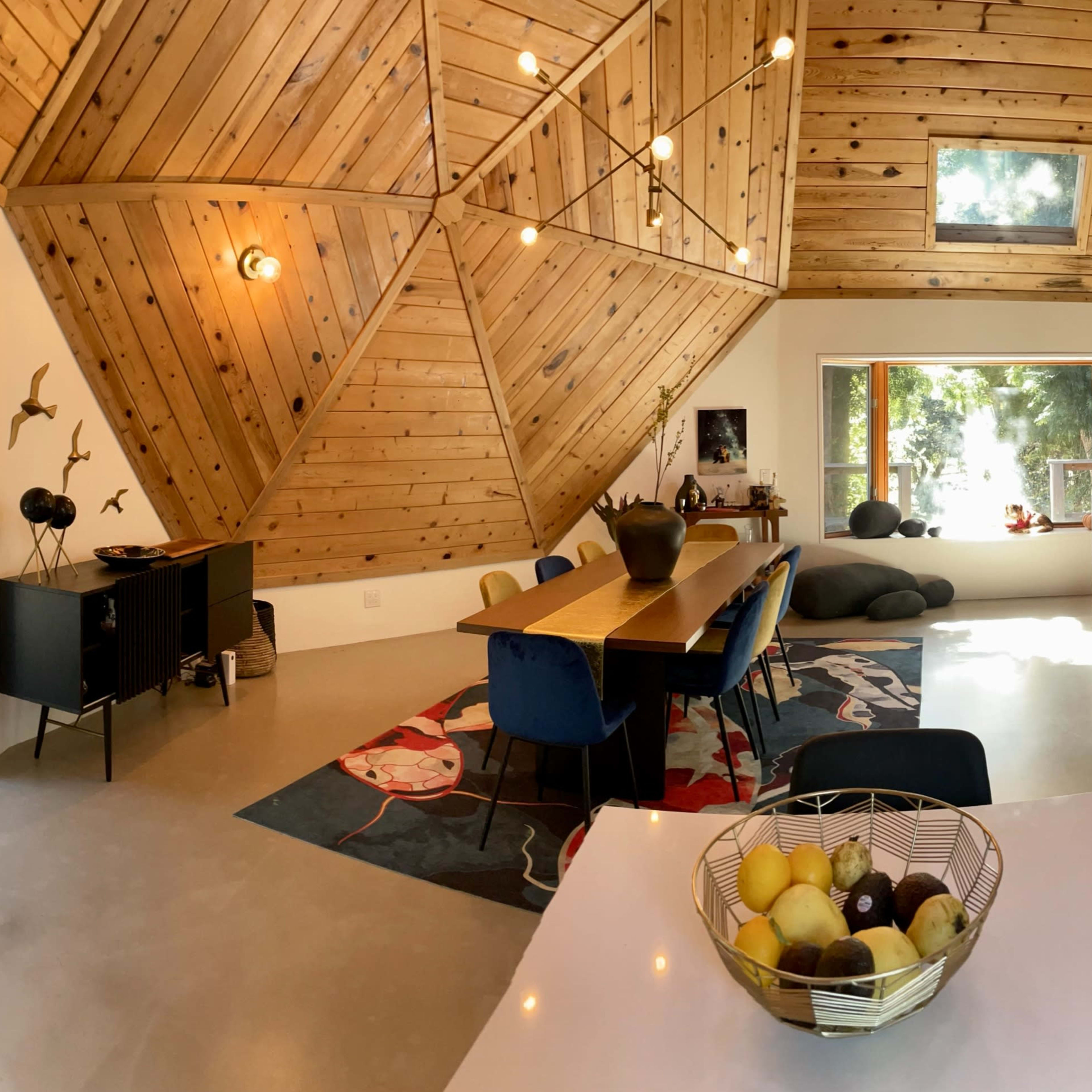 A modern, triangular-shaped interior space featuring a wooden ceiling, a long dining table with blue chairs, and a kitchen area with a basket of fruit on the counter.