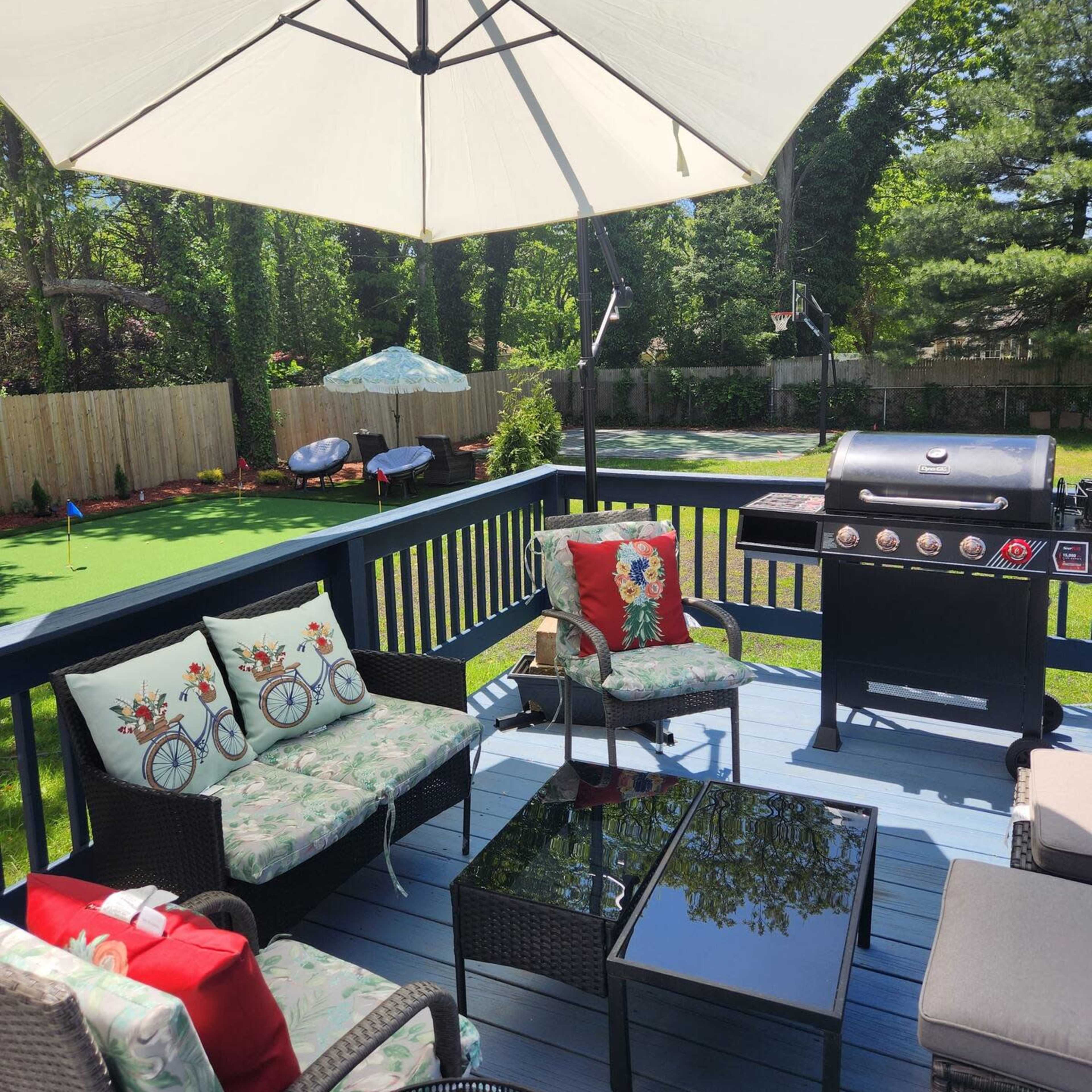 A backyard deck area with a grill, outdoor seating, and a large umbrella, surrounded by a lawn and trees.