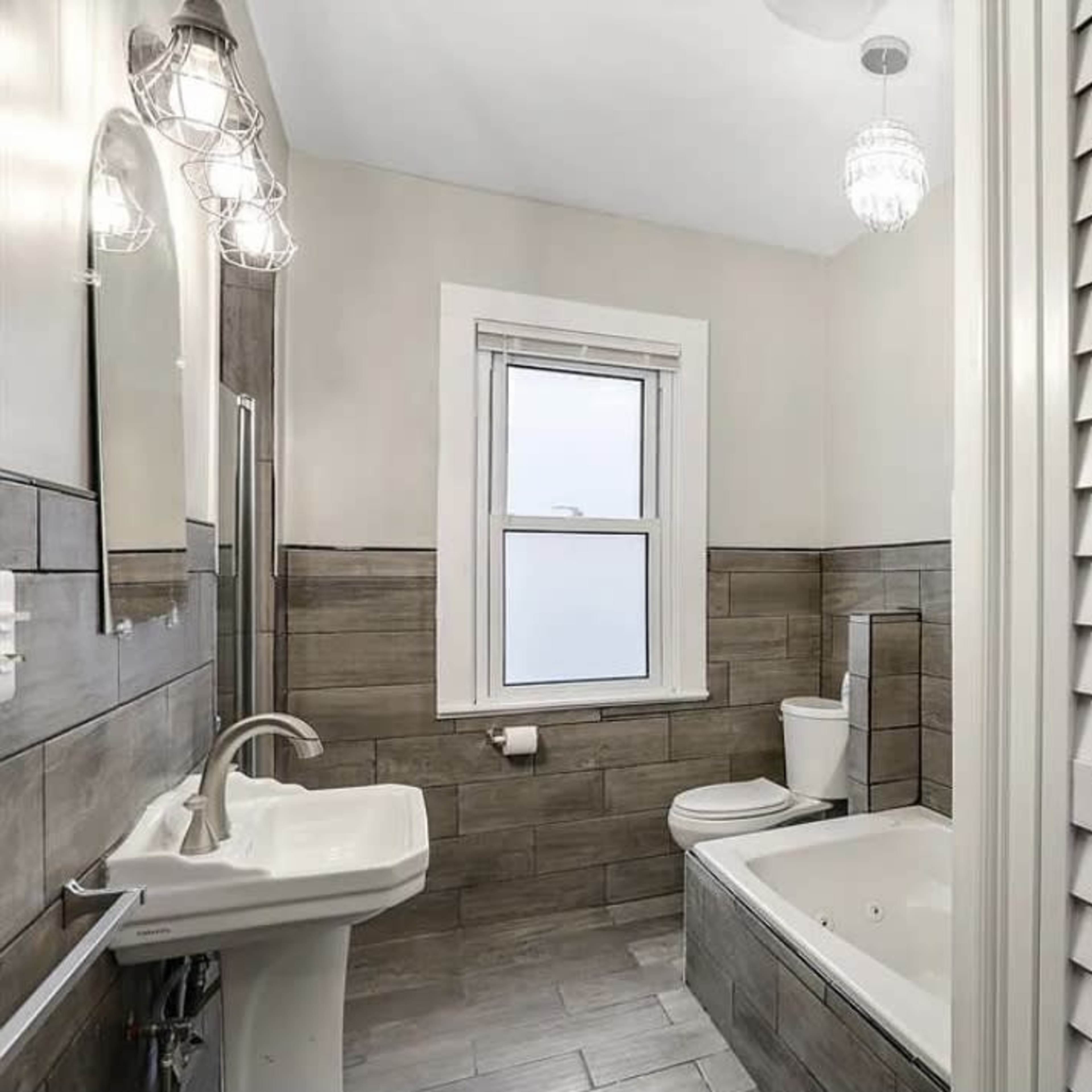 A modern bathroom with a bathtub, a pedestal sink, a toilet, and a window, featuring tiled walls and a light fixture.